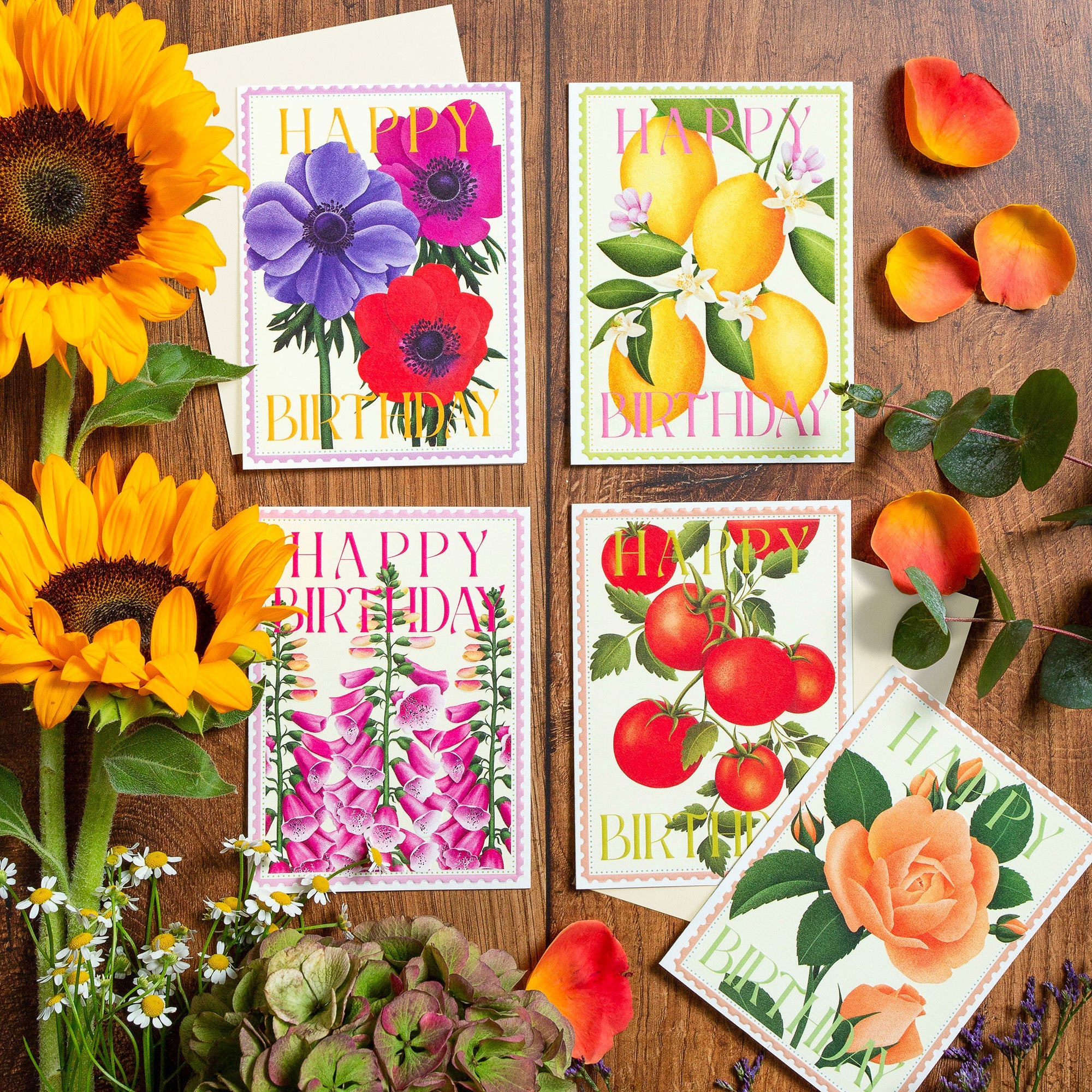 A collection of beautiful botanical birthday cards called 'Colourburst', laid out with fresh flowers on a wooden table.