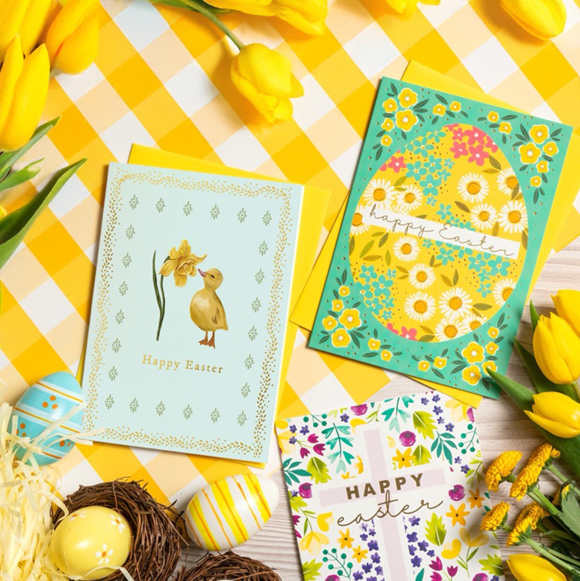 A collection of Easter cards are laid out with Easter eggs, fresh flowers and a tablecloth.
