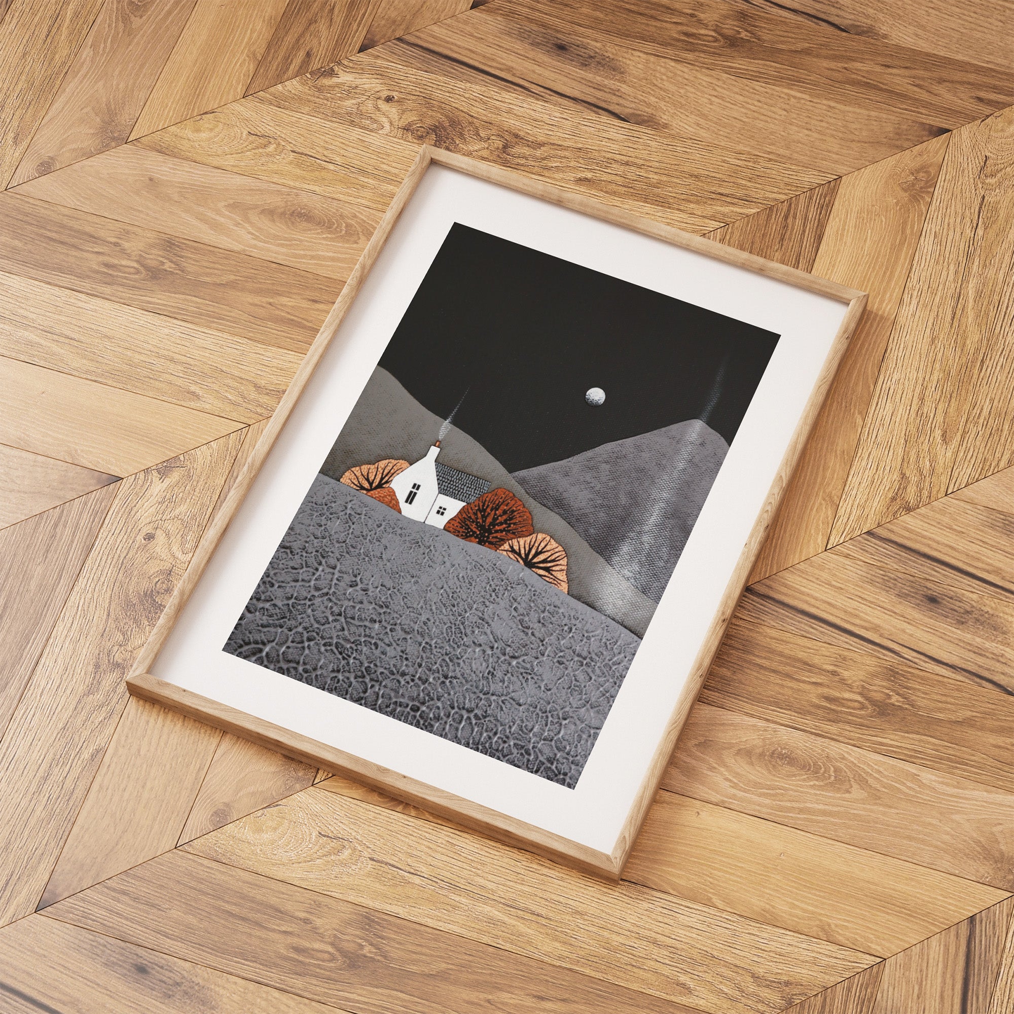 Framed artwork of a house on a mountain with a moonlit sky on a wooden floor.