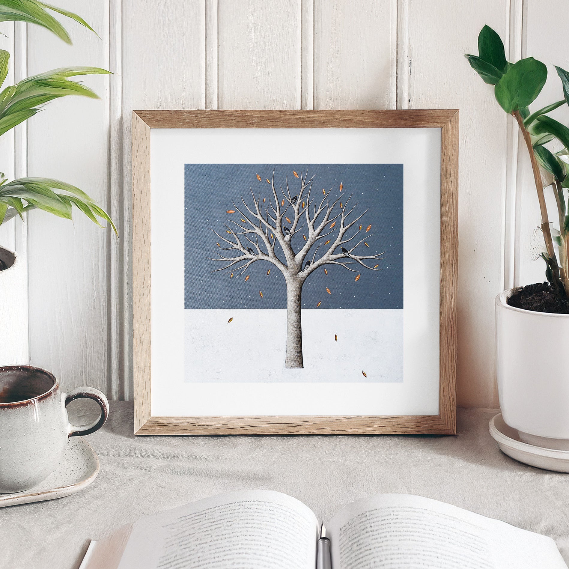 Framed artwork of a tree with birds on a blue background, placed on a surface with plants and a cup.