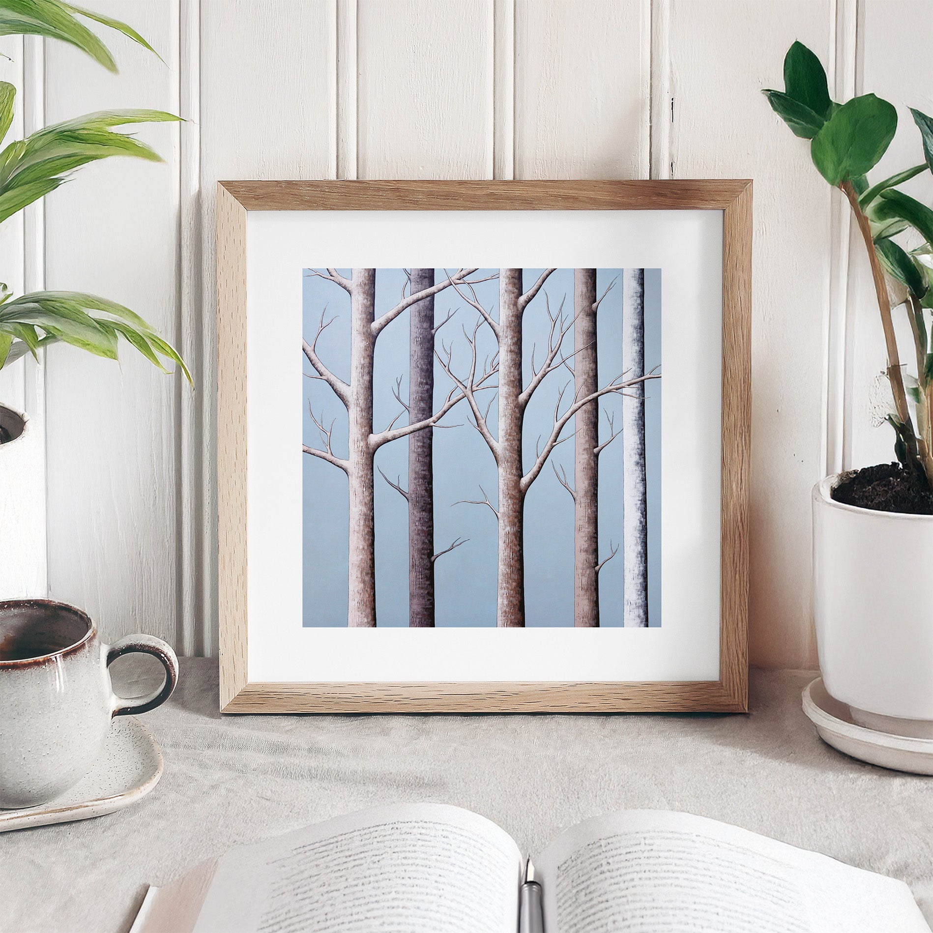 Framed artwork of trees on a table with plants and a cup in the background.