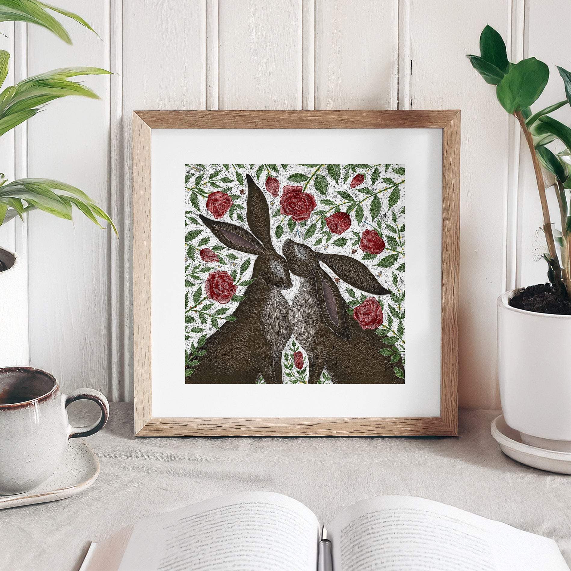 Framed artwork of two rabbits among roses on a table with plants and a cup.