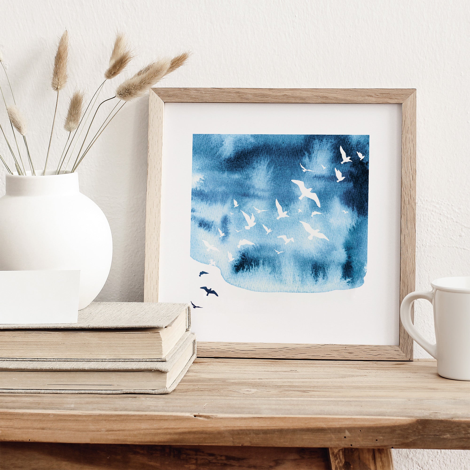 Framed artwork of birds on a blue watercolor background on a wooden surface with decor items.