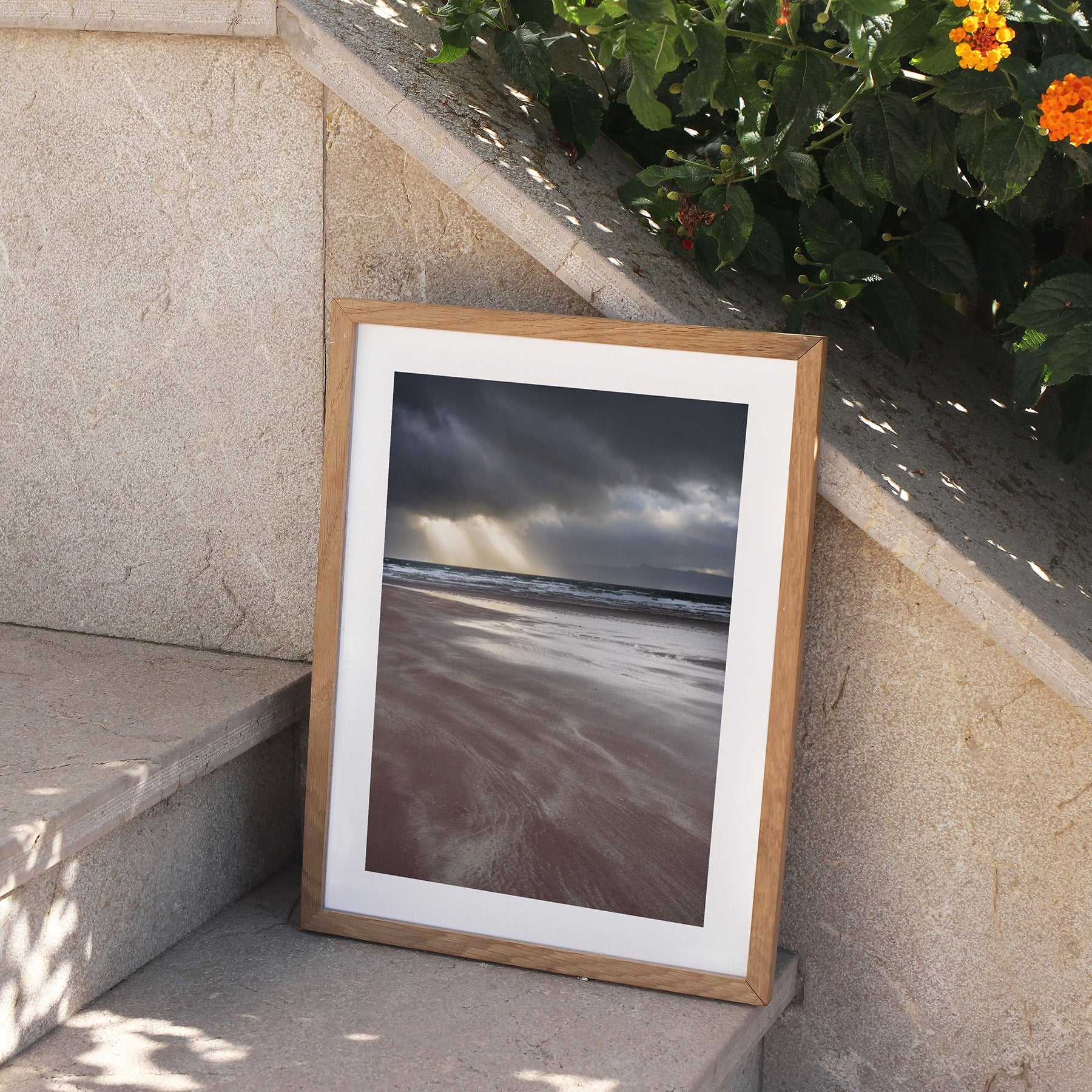 Framed photography art print featuring a coastal view of the the beach and the sea, with beams of light peeking through moody clouds. Framed print is perching on stone steps, outside.