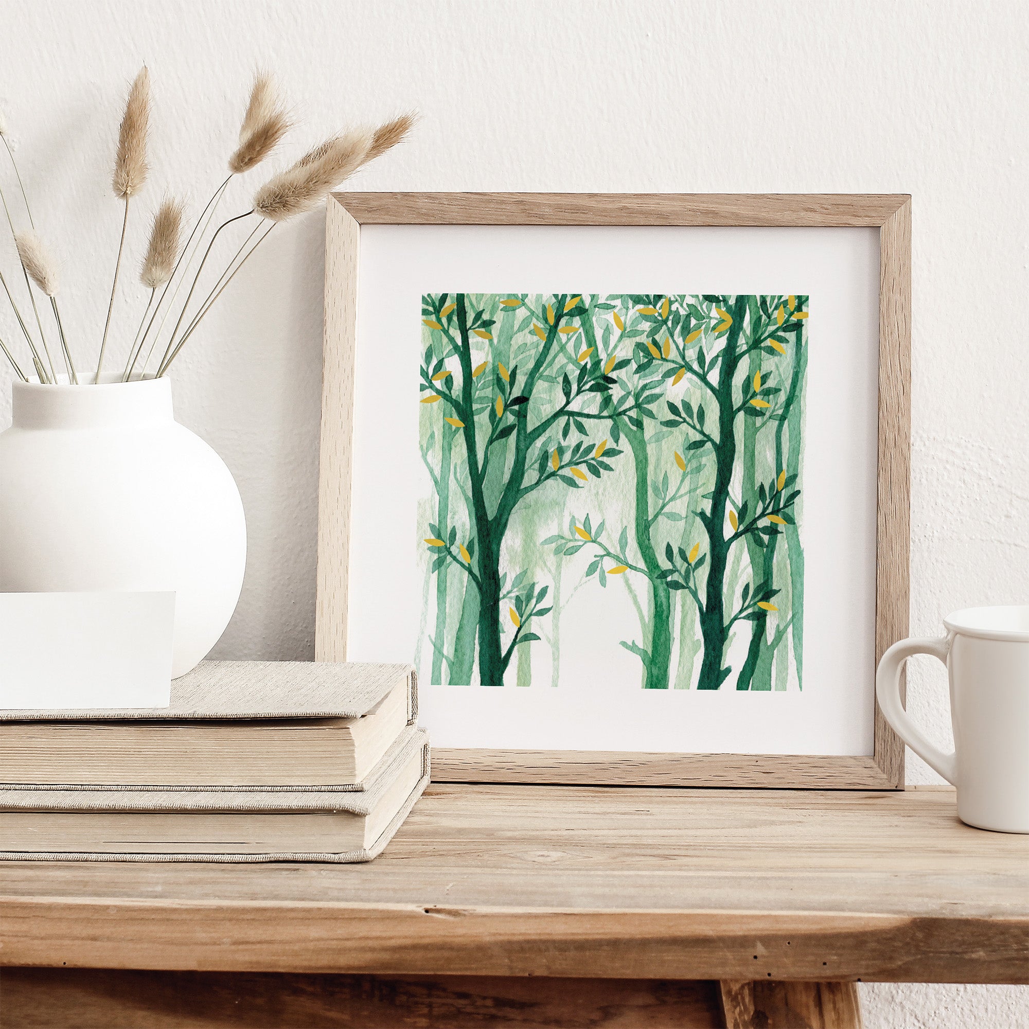 Framed artwork of green trees on a wooden surface with books and a mug.
