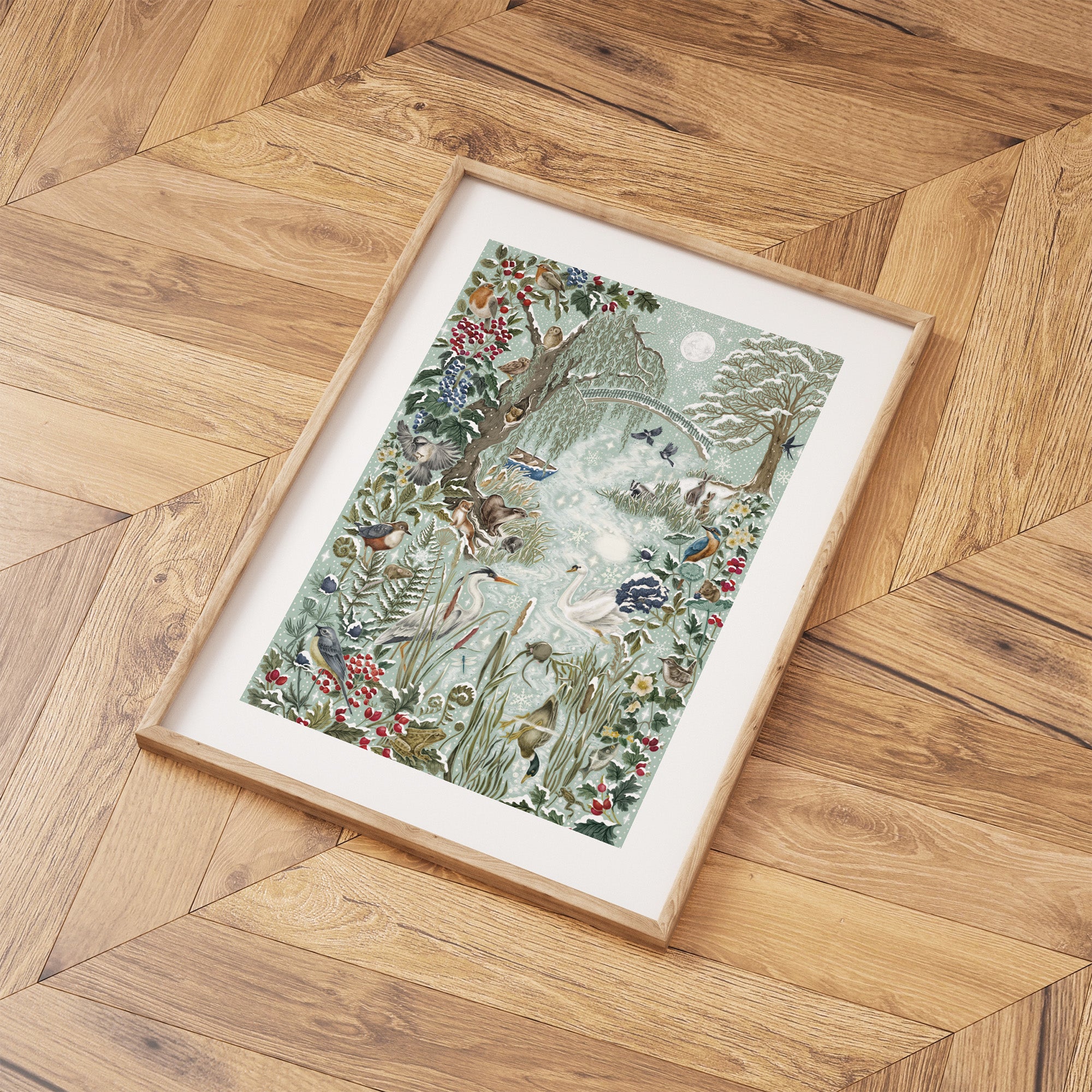 Framed Christmas art print featuring a wintery wonderland scene of animals and botanicals basking by a river on a frosty day. Print is laid out on a wooden floor.