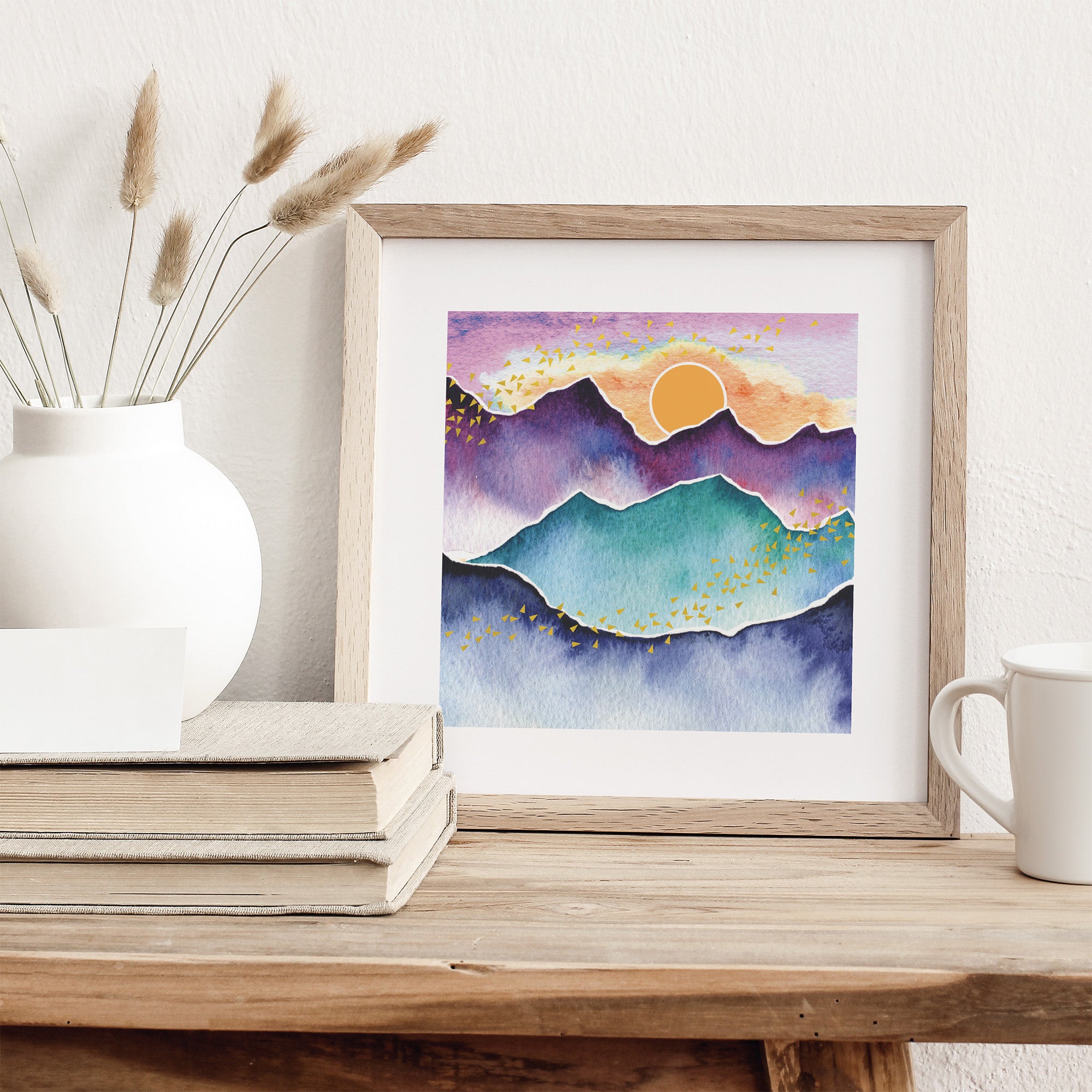 Framed artwork of mountains and sun on a wooden surface with decor items.