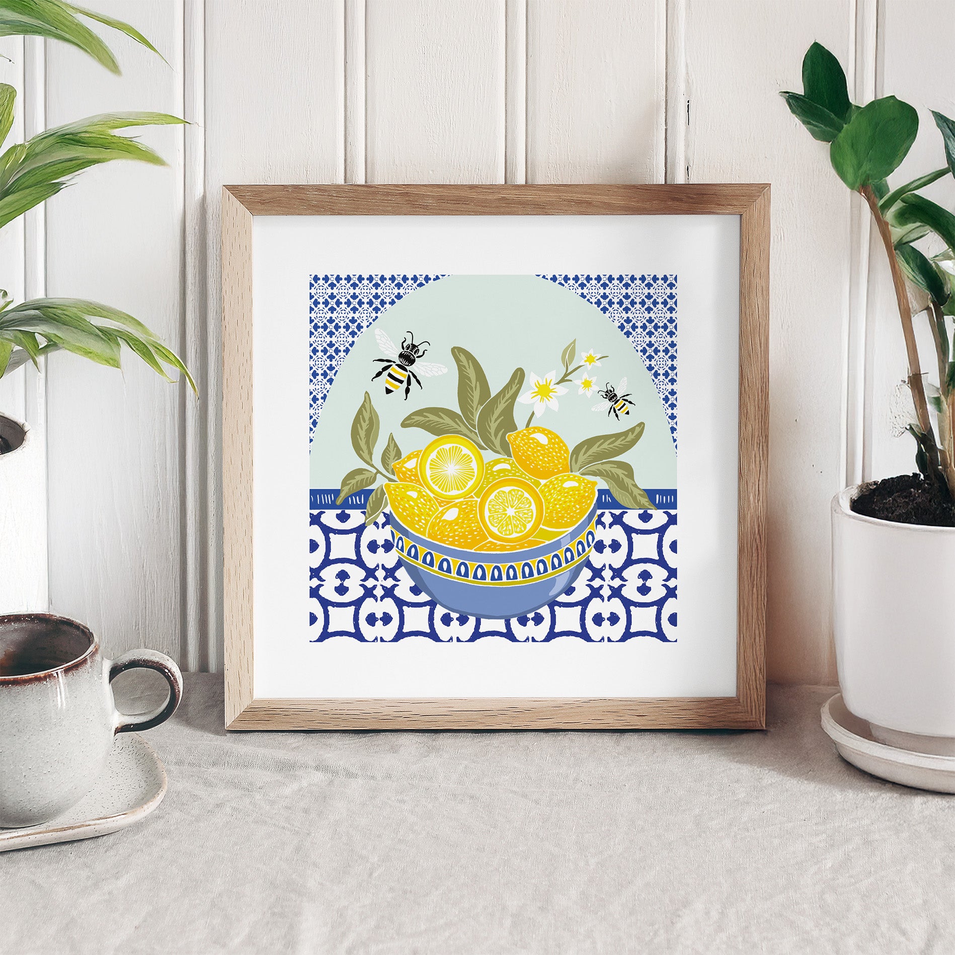 Framed artwork with a bowl of lemons and bees on a decorative pattern, placed on a surface with plants and a cup.