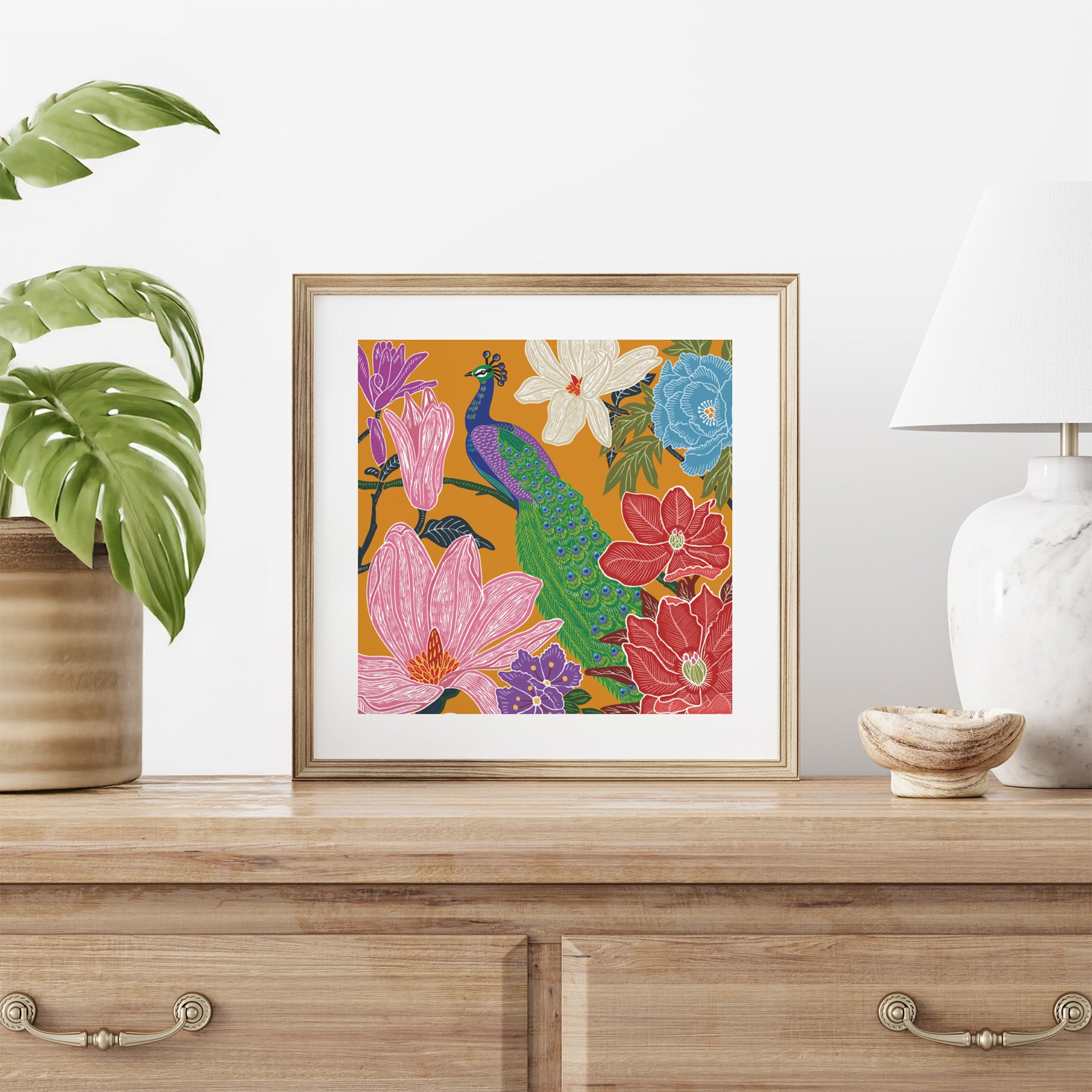 Framed colorful floral and peacock artwork on a wooden surface with decorative items.