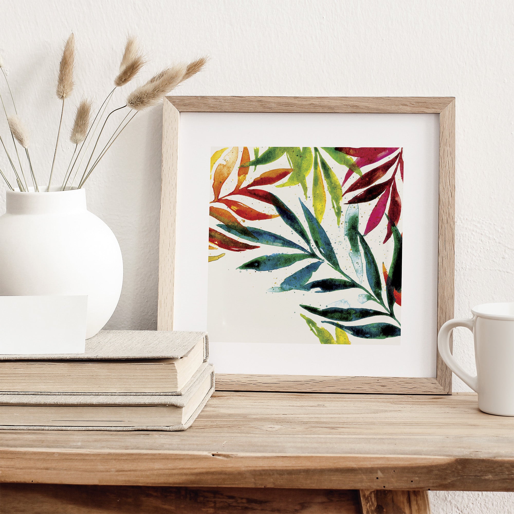 A watercolour art print of colourful leaves. Art print is framed and sits on a dresser.