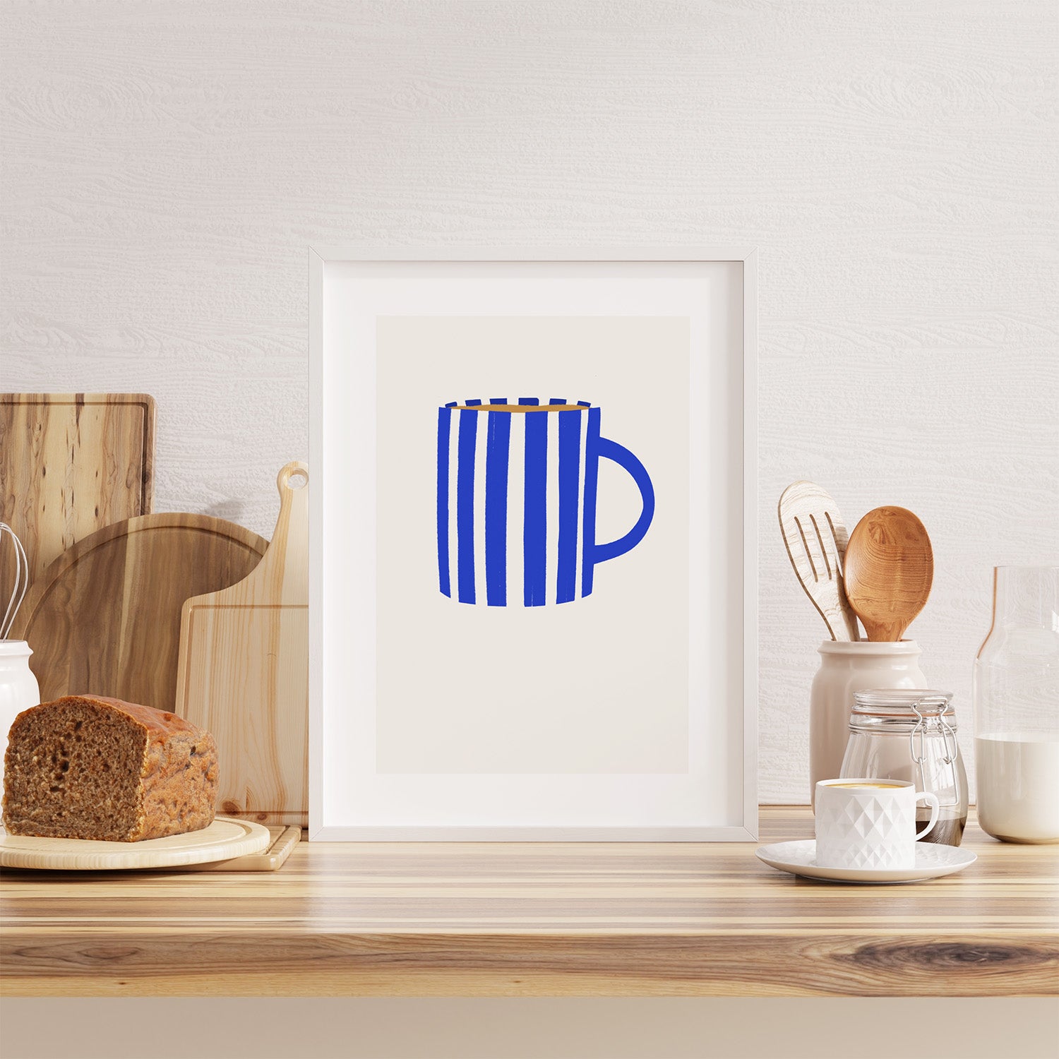 Framed artwork of a blue mug on a kitchen counter with utensils and bread.