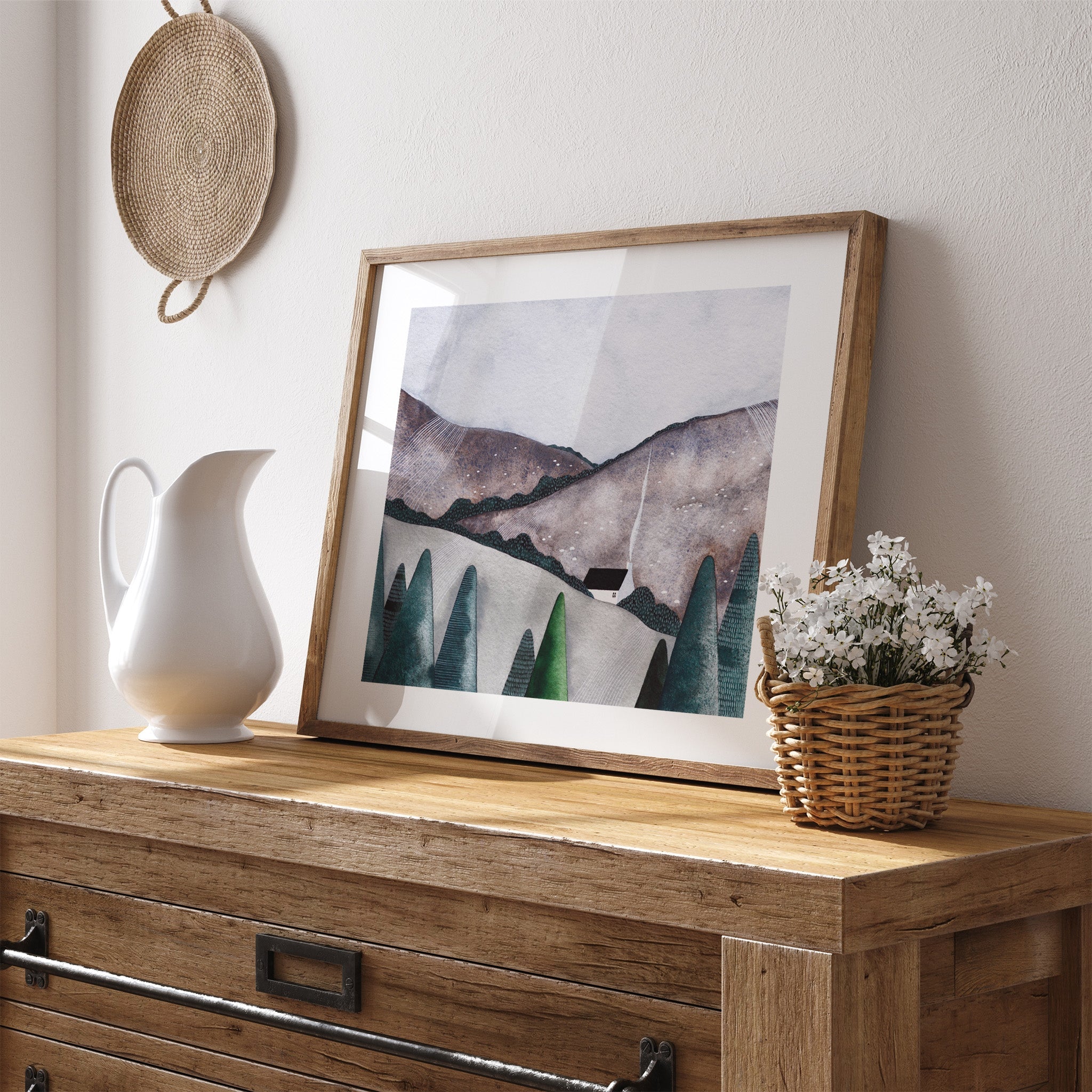 Art print featuring a white house nestled in the countryside, surrounded by green forest. Art print is leaning against a white wall near flowers.