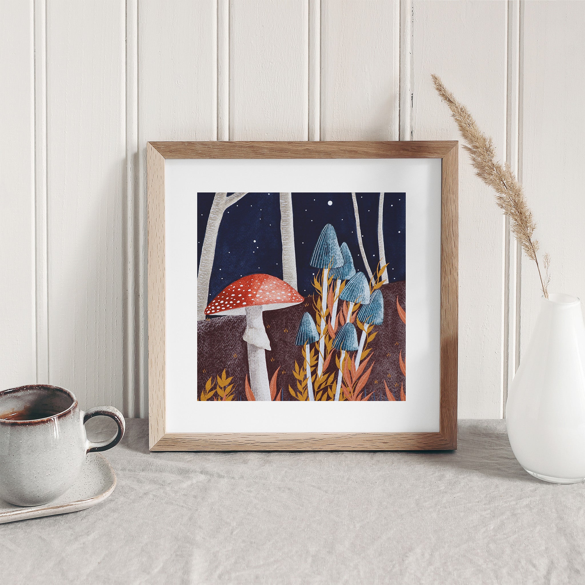 Art print of mushrooms growing in a dark Autumnal forest. Art print is framed and leaning against a white wall.
