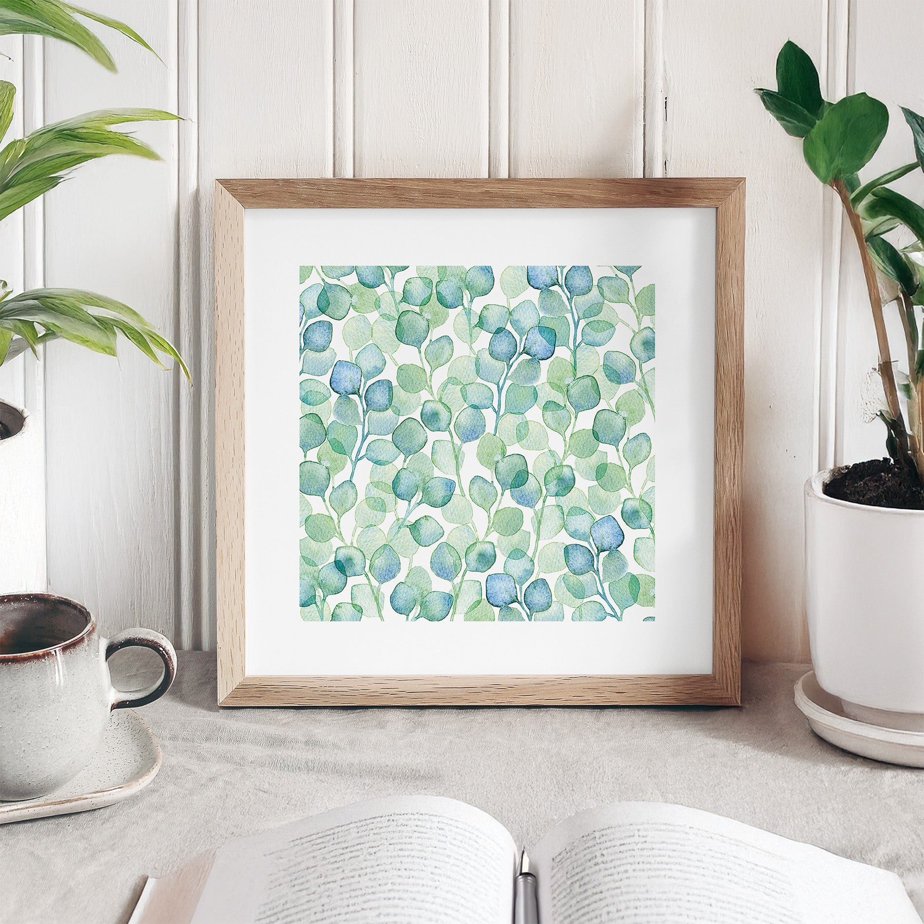 Framed artwork with green and blue leaf pattern on a white surface with plants and a cup.