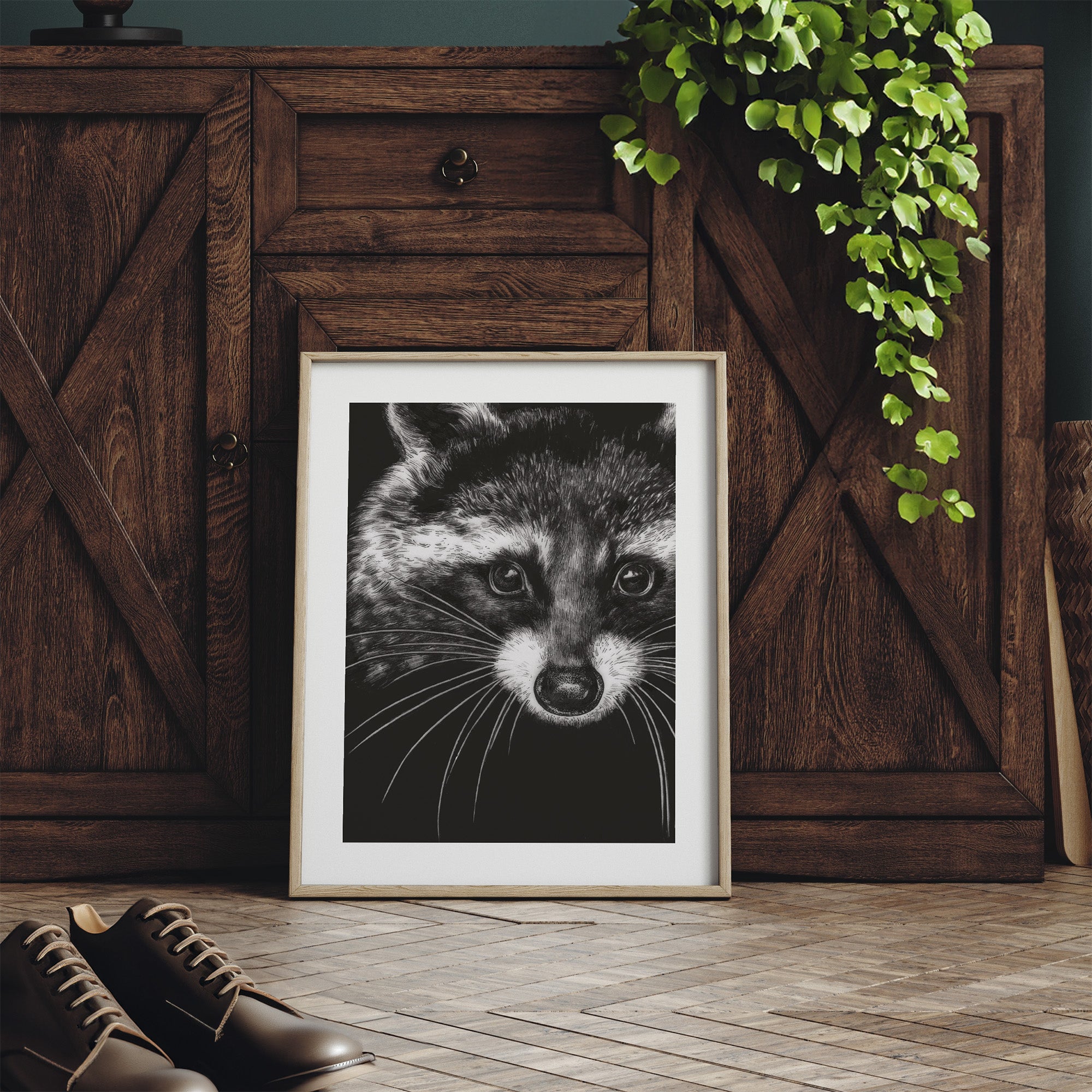 Framed black and white raccoon print on a wooden floor with a wooden cabinet and plant in the background.