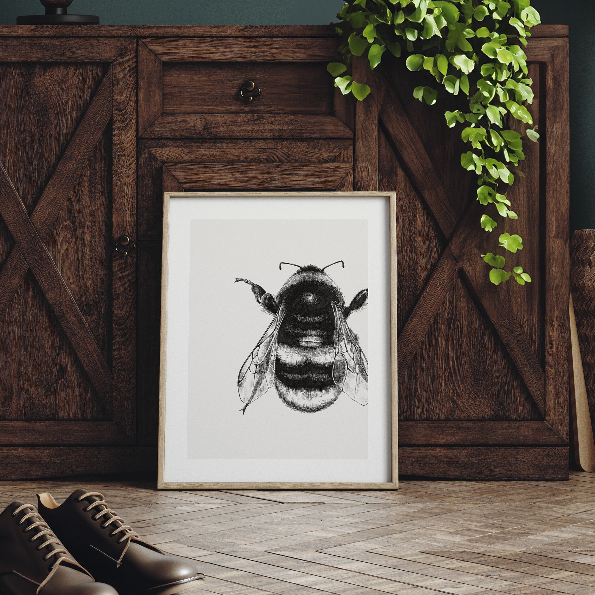 Framed black and white bee illustration on a wooden floor with a wooden cabinet and plant in the background.