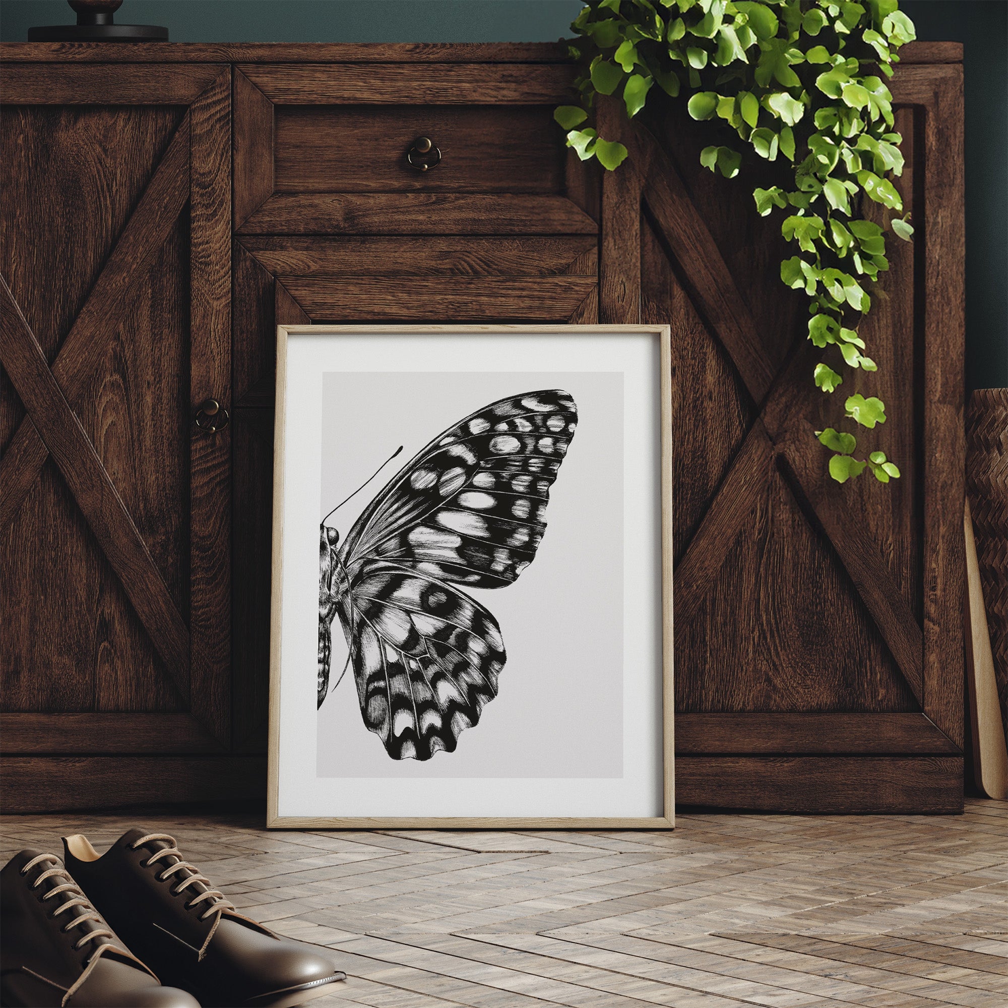 Framed butterfly artwork on a wooden floor with a wooden cabinet and plant in the background.