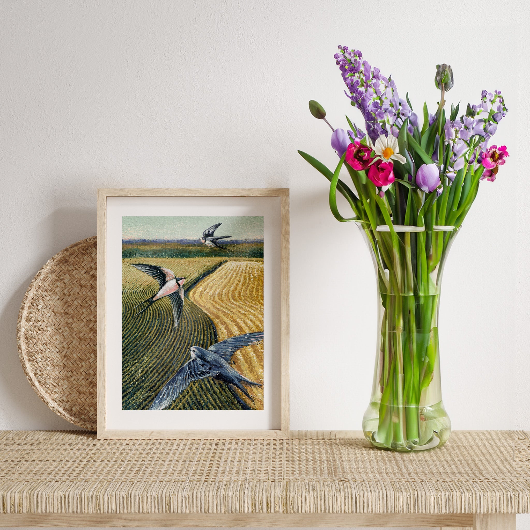 A watercolour art print of three swallows flying above farmer's fields. The print is framed on a tablet next to a vase of flowers.