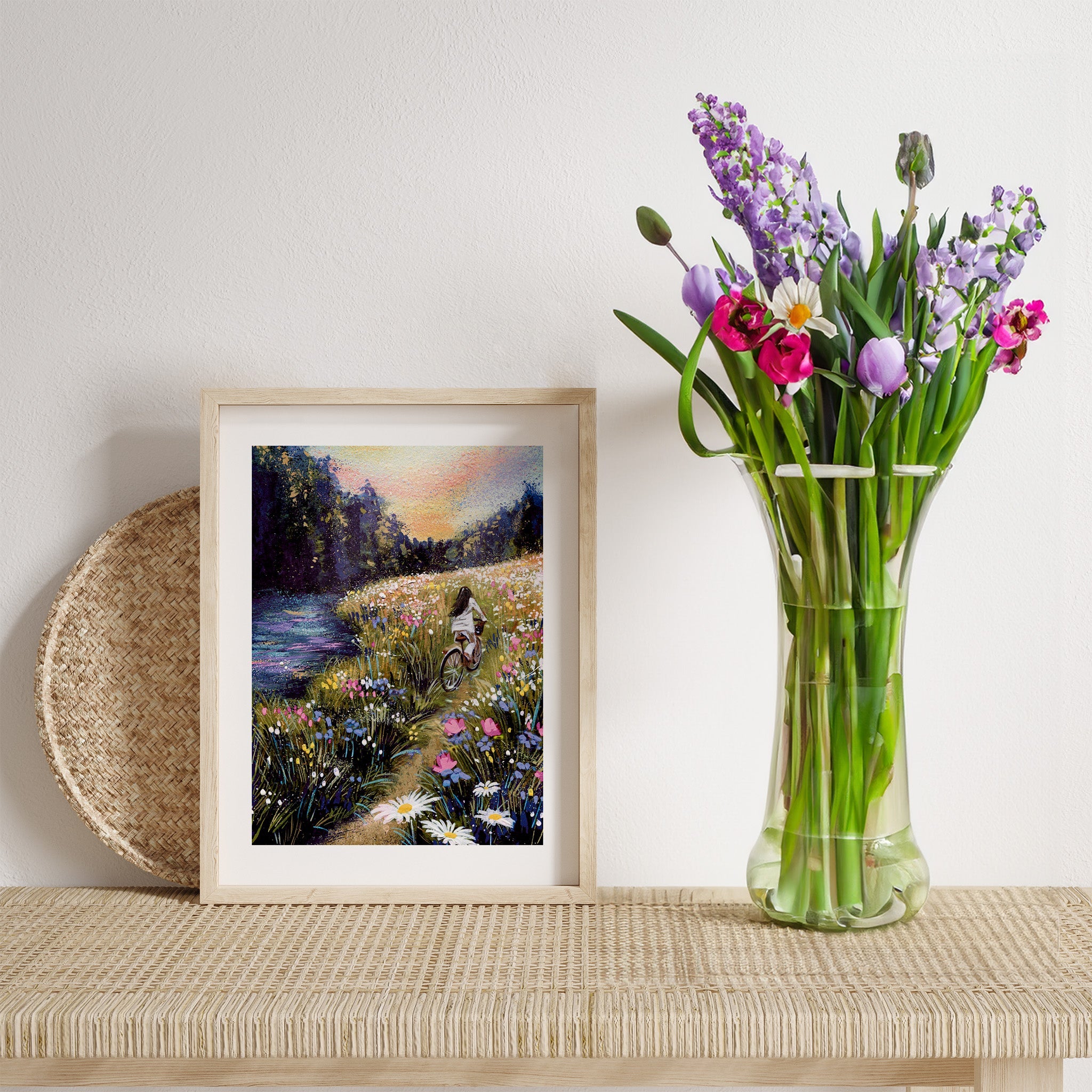 A print of a woman riding a bike through a Summer forest, framed and displayed on a table next to a vase of flowers.