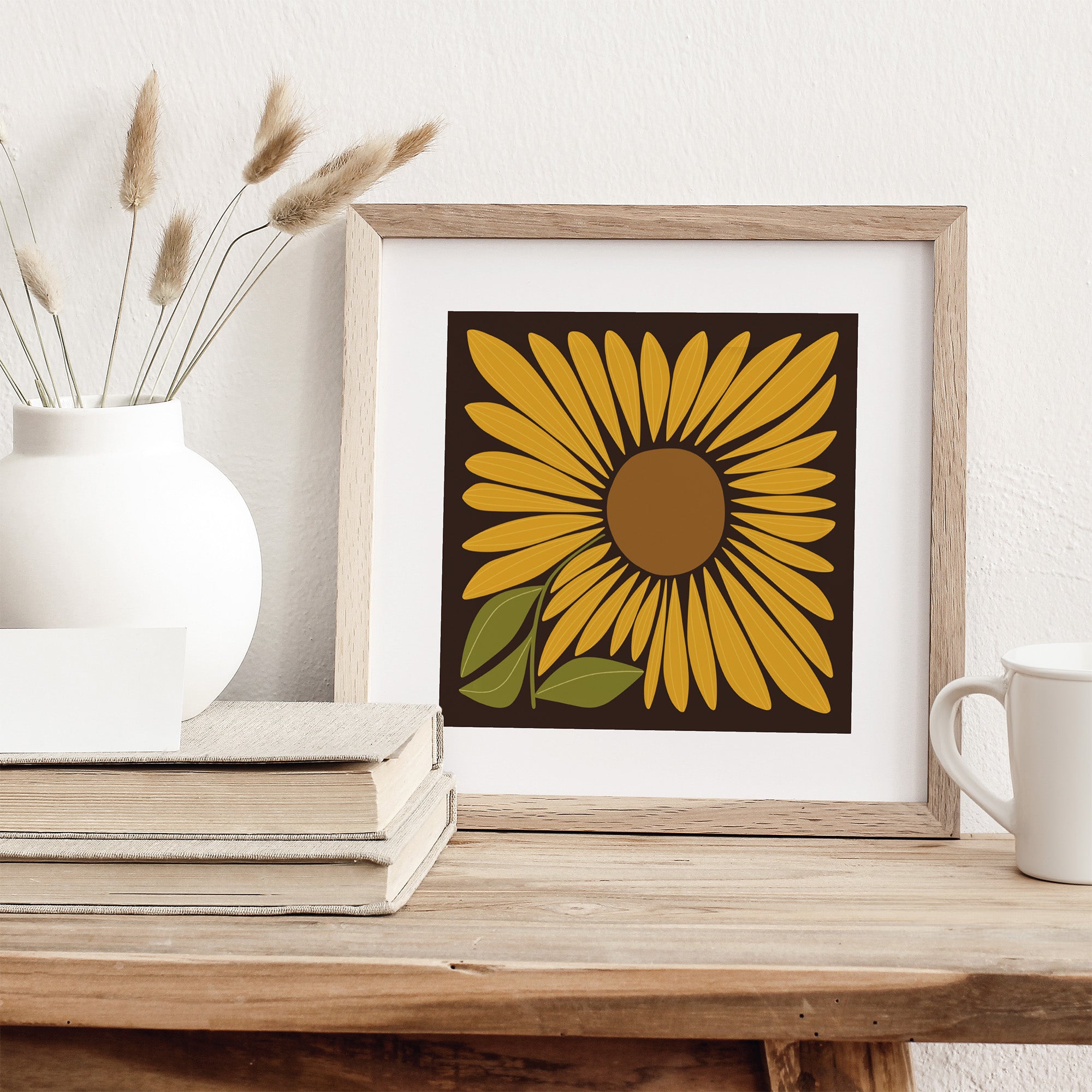 Framed sunflower artwork on a wooden surface with a white vase and books.