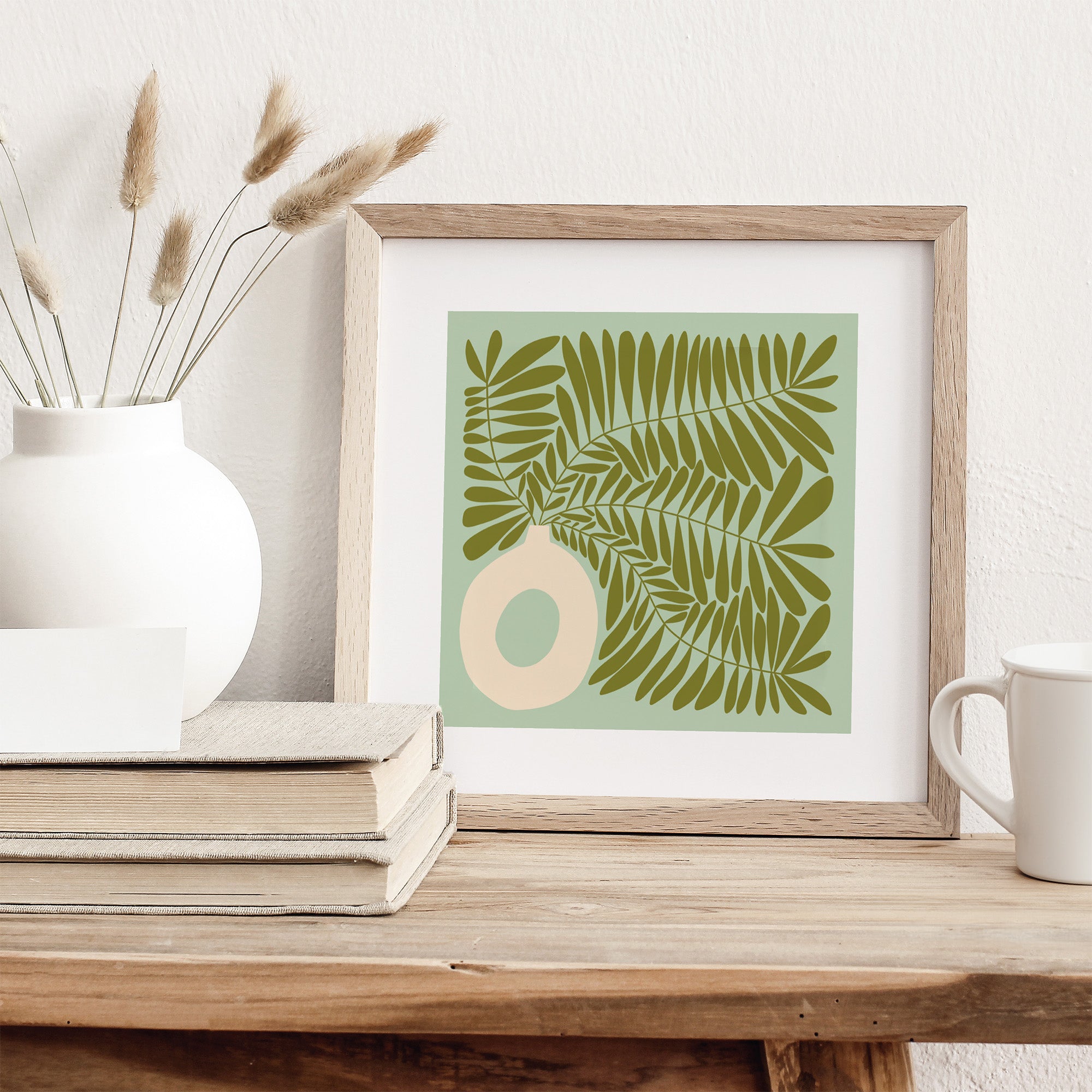 Framed artwork with green leaf design on a wooden surface with books and a vase.