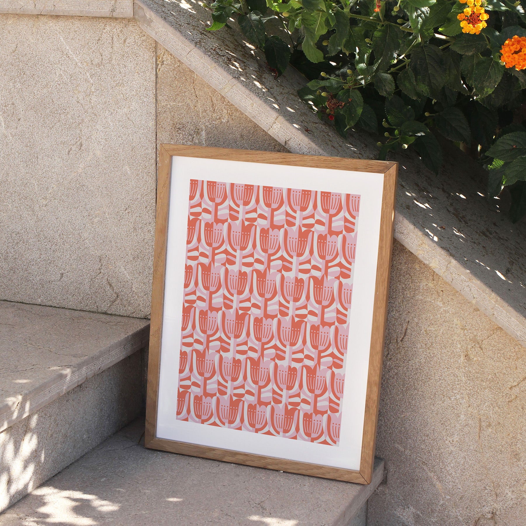 Framed artwork with a pink geometric pattern on a white background, leaning against a stone wall with plants.