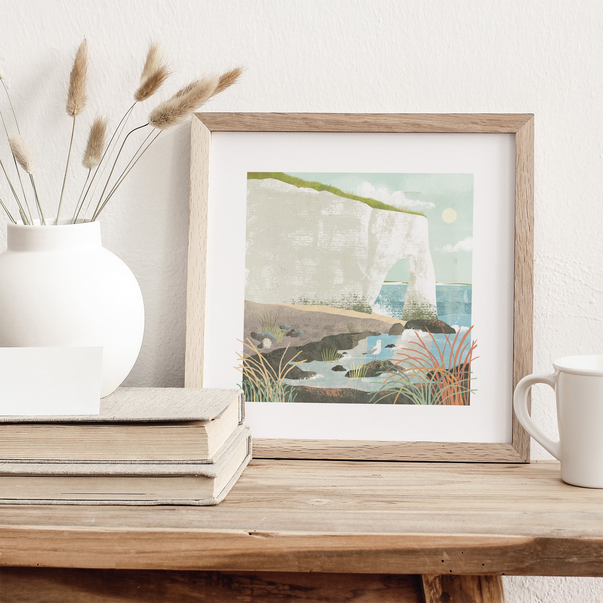 Framed artwork of a coastal scene on a wooden surface with a vase and books.
