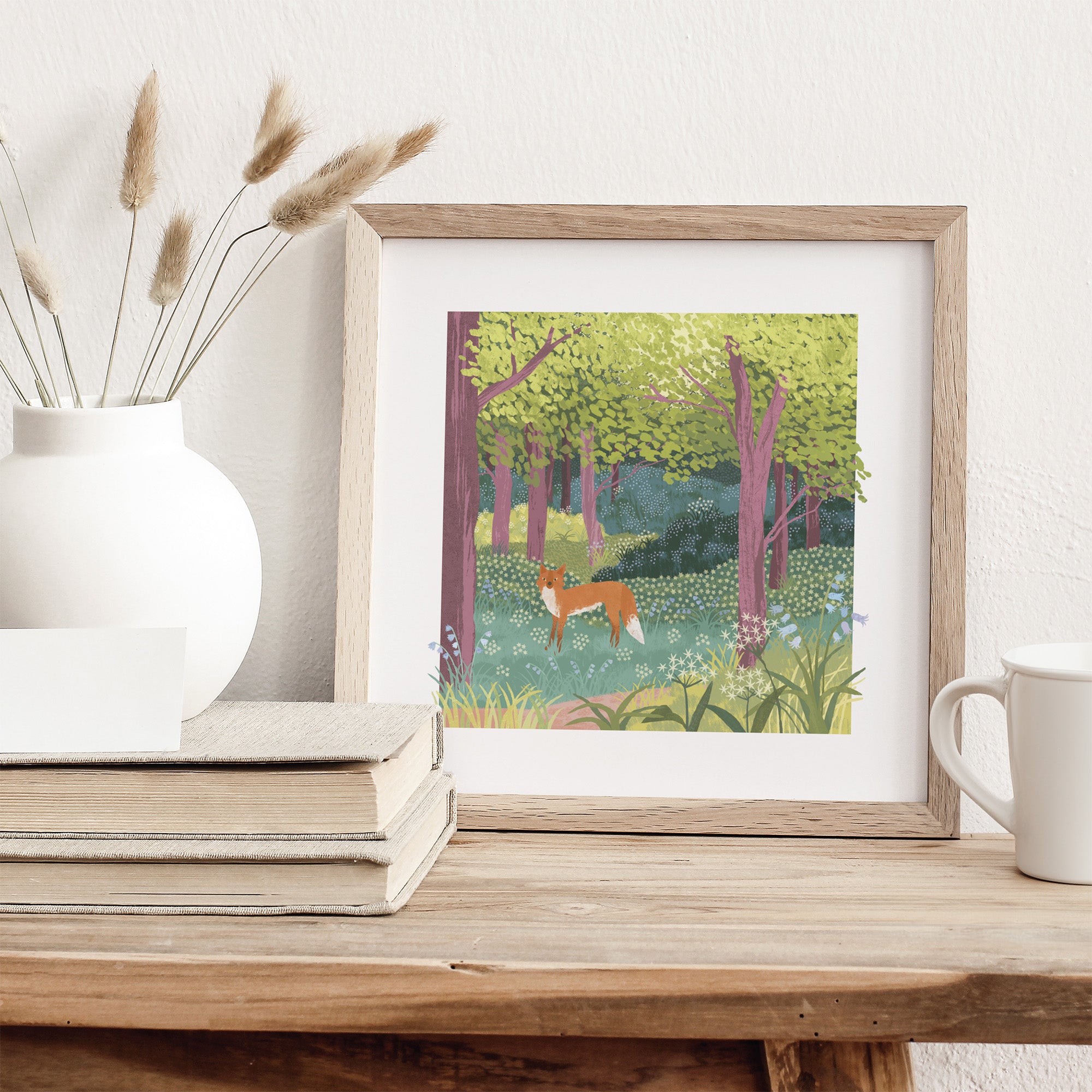 Framed artwork of a fox in a forest on a wooden surface with a white vase and books.