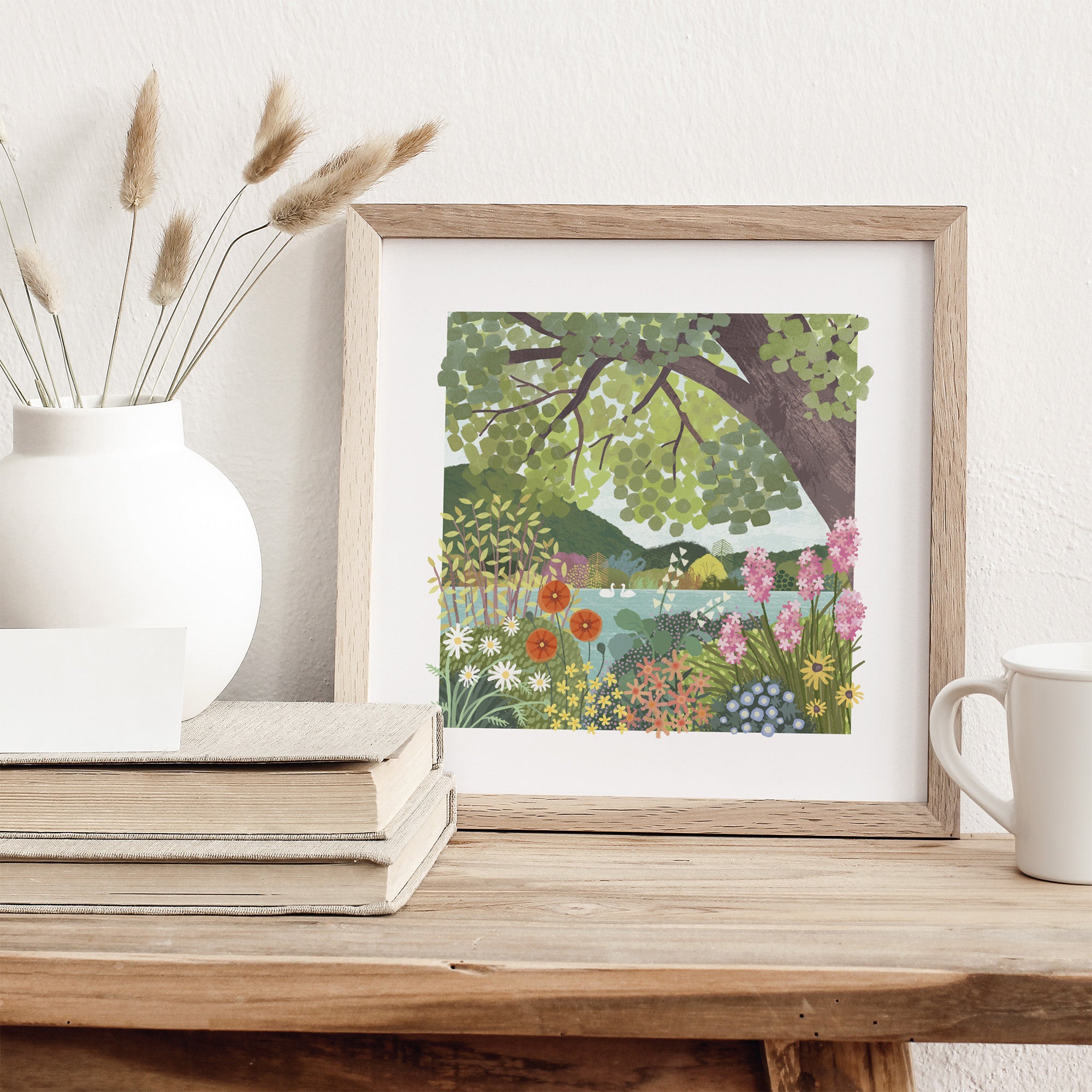 Framed artwork of a nature scene on a wooden surface with a vase and mug.