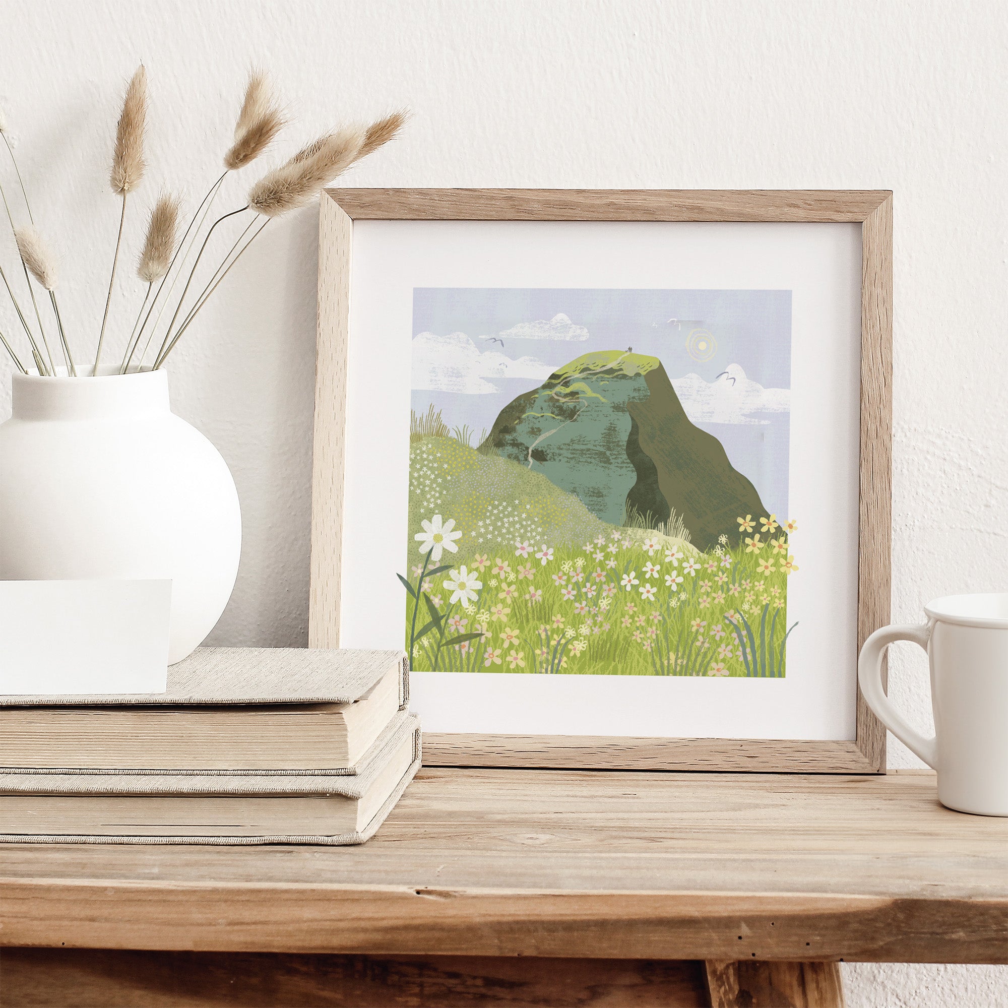 Framed artwork of a mountain landscape on a wooden surface with a white vase and books.