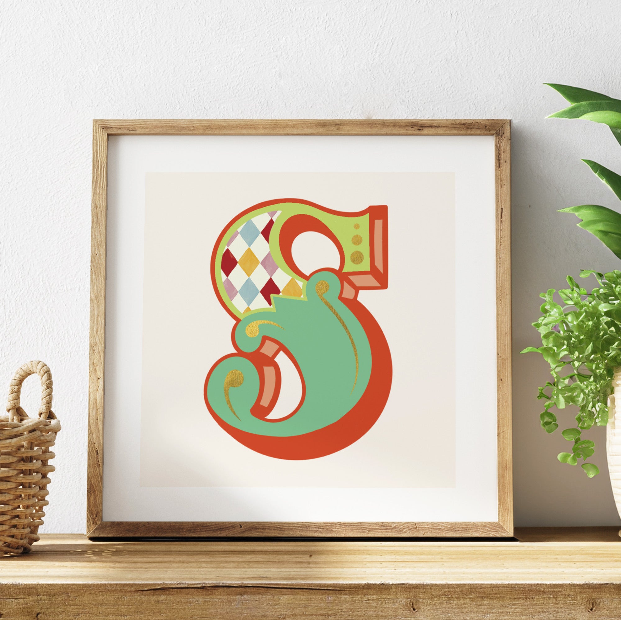 Framed decorative letter 'S' on a shelf with a plant in the background.
