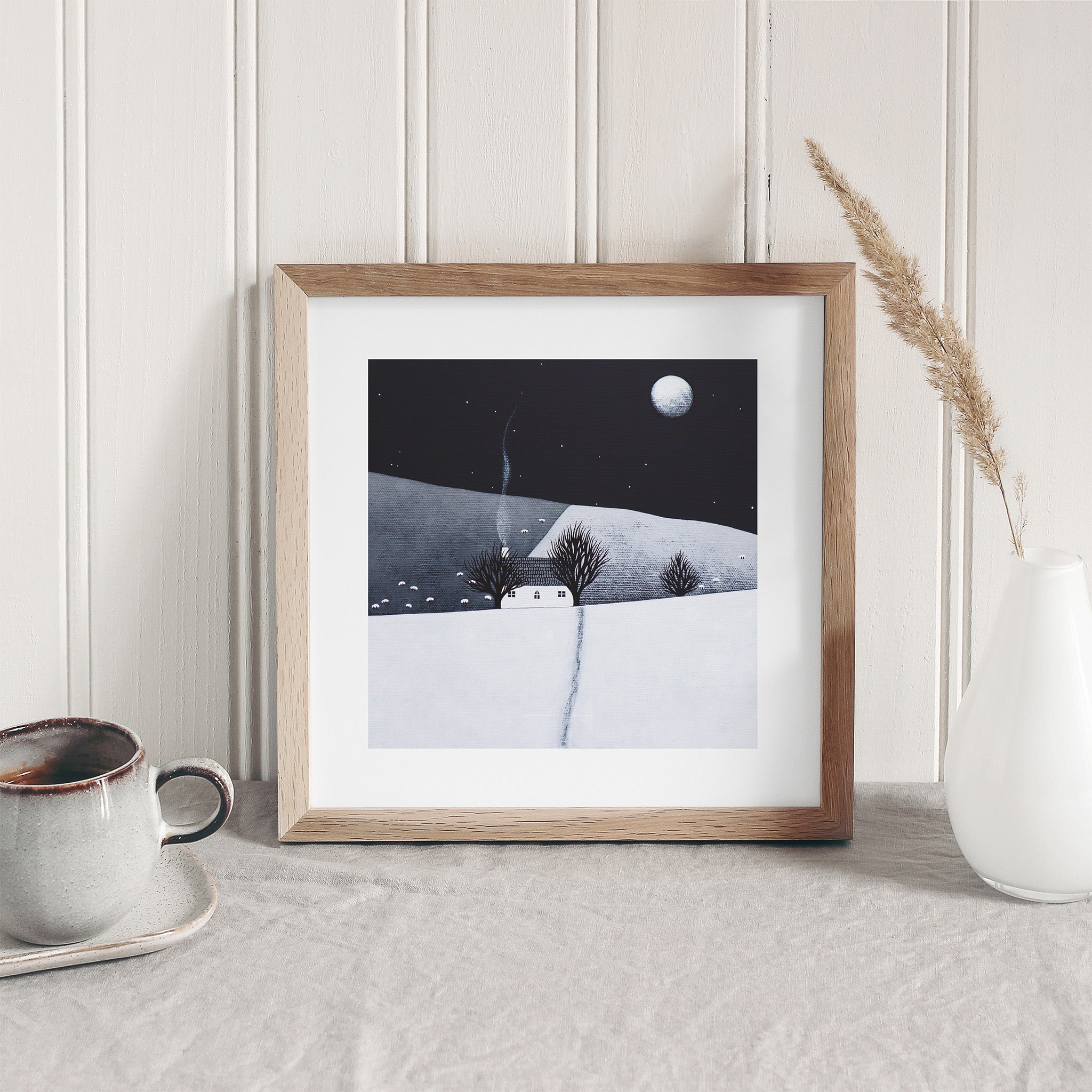 Art print of a cosy home in a wintery moonlit field. Art print is leaning against a white wall.