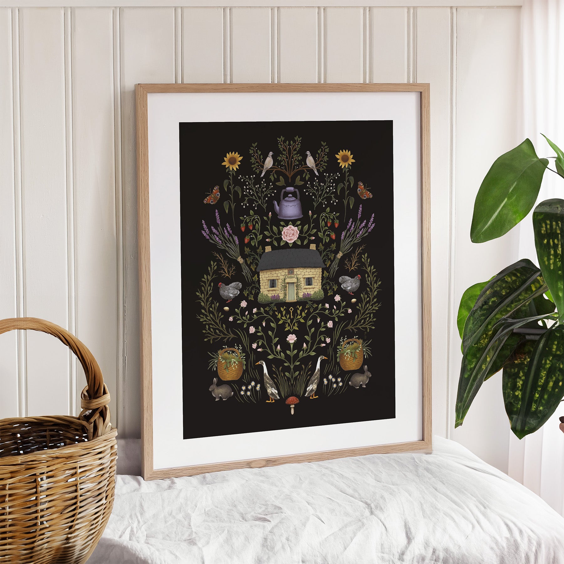Framed art print featuring a cottage surrounded by botanicals and wildlife on a black background. Art print is leaning against a wall near a plant.