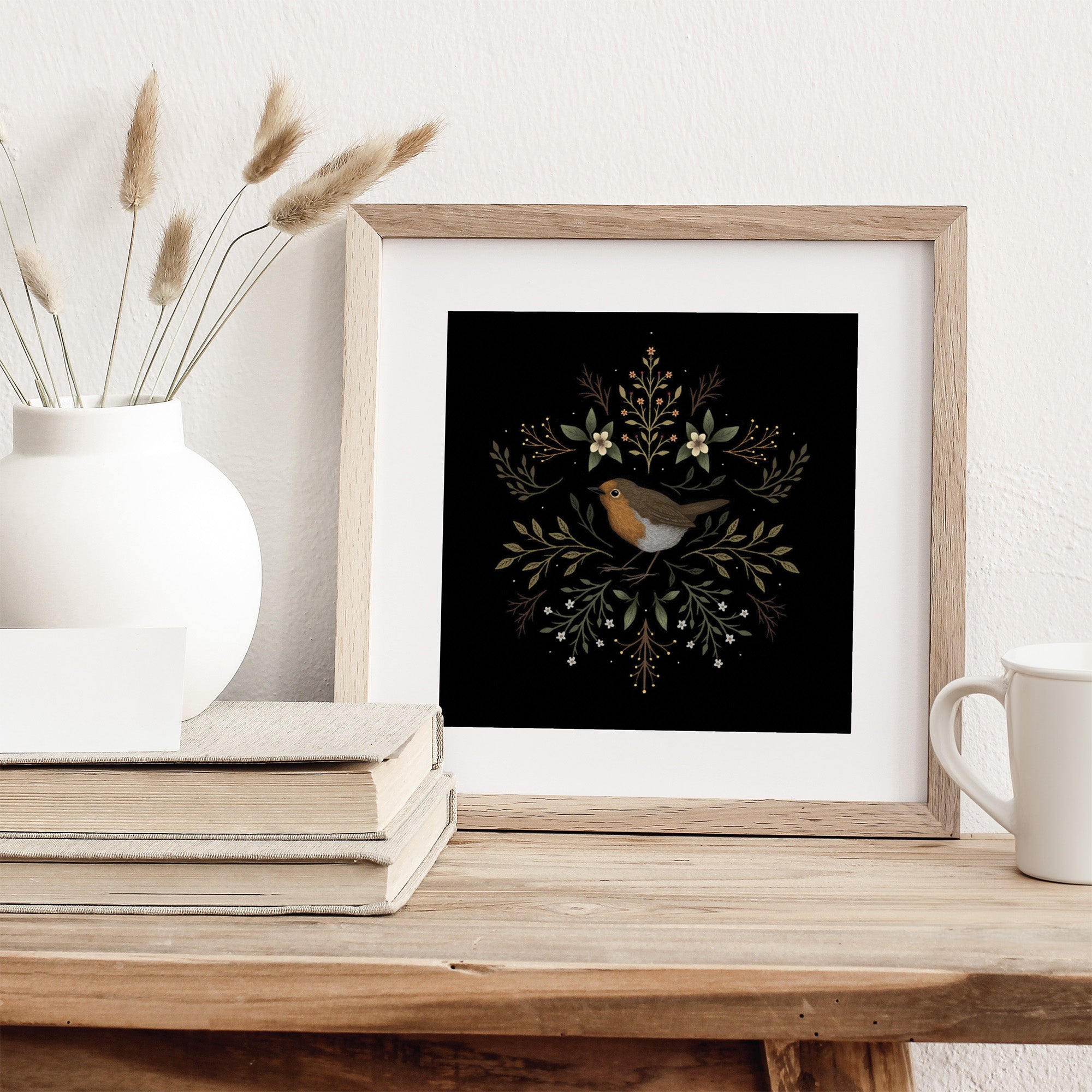 Framed art print featuring a British robin surrounded by botanicals on a black background. Print is leaning against a white wall on a table.