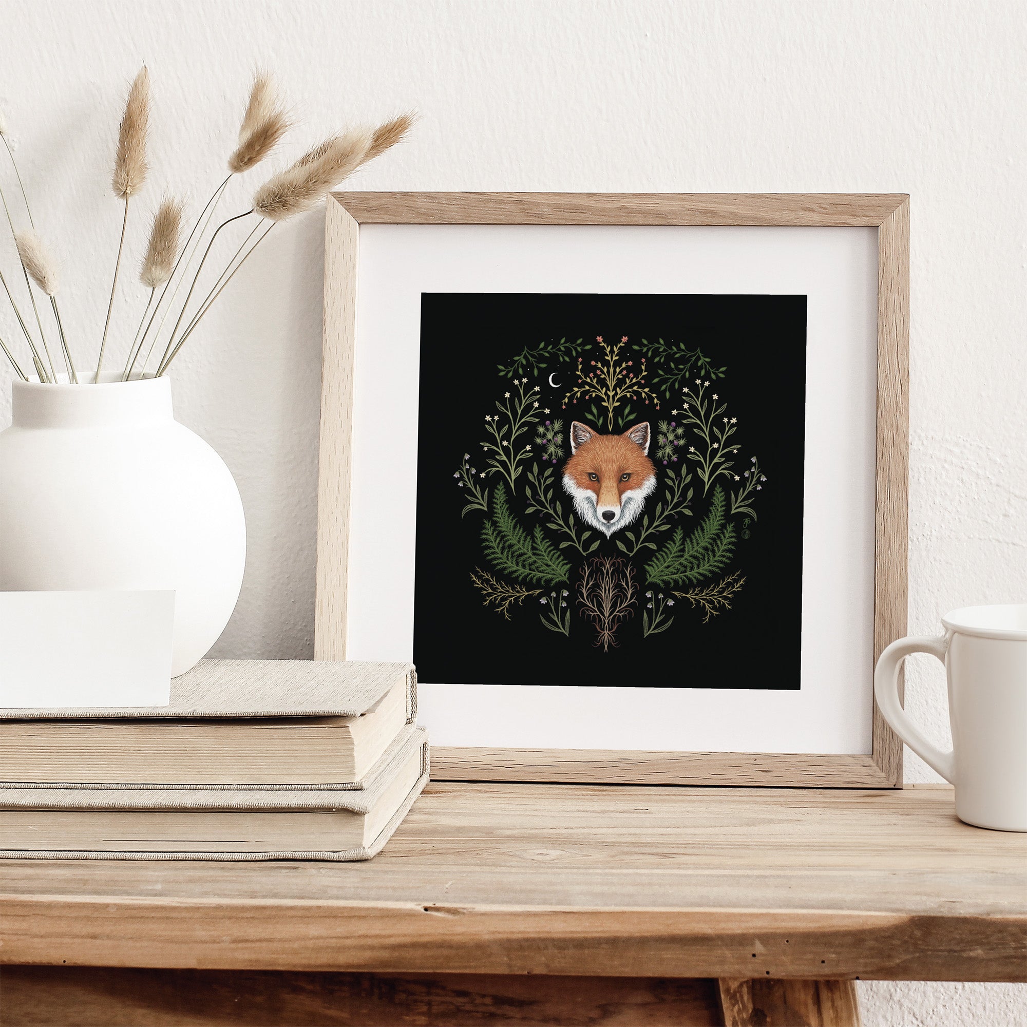 Framed art print featuring a British fox surrounded by botanicals on a black background. Print is leaning against a wall.