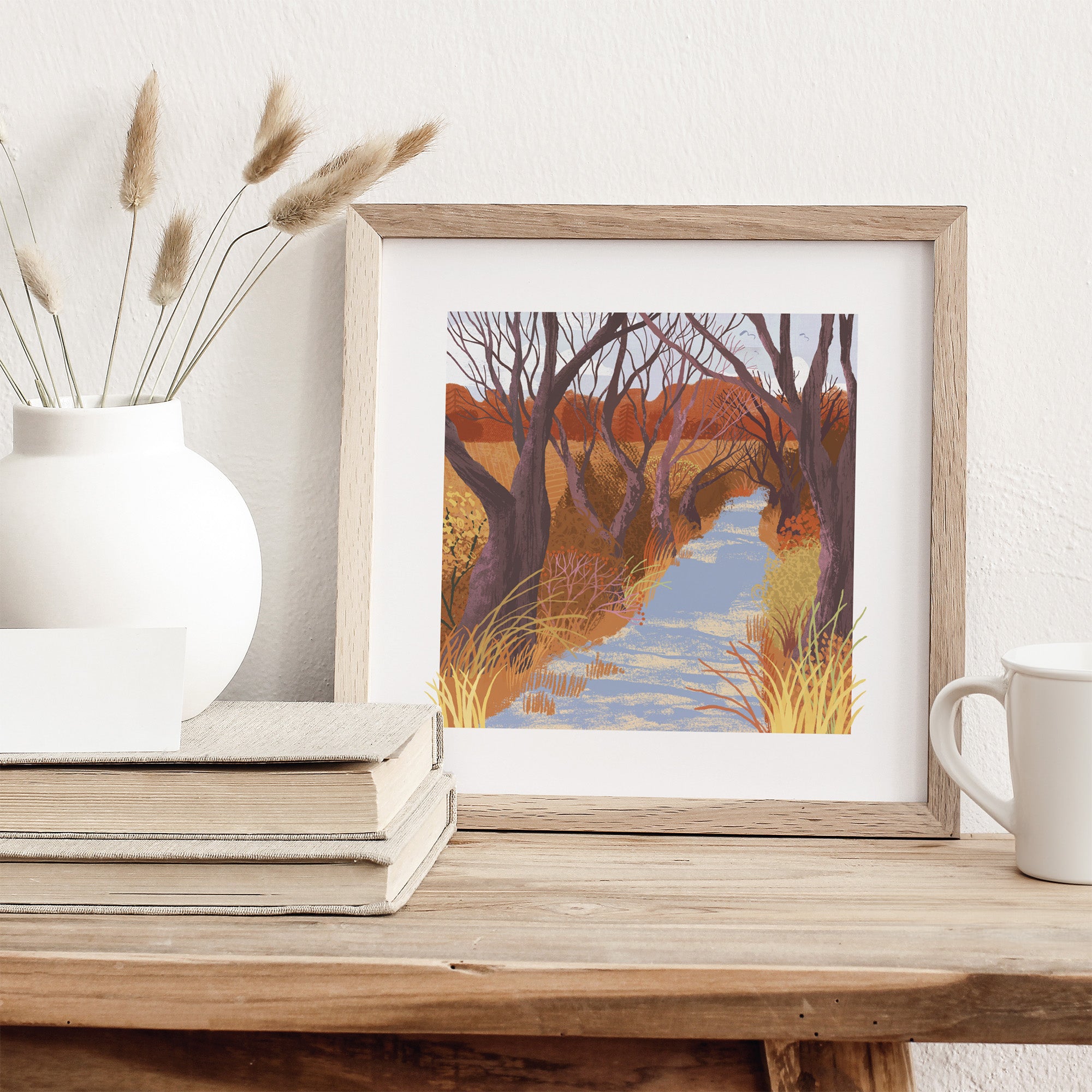 Framed artwork of a nature scene on a wooden surface with a vase and books.