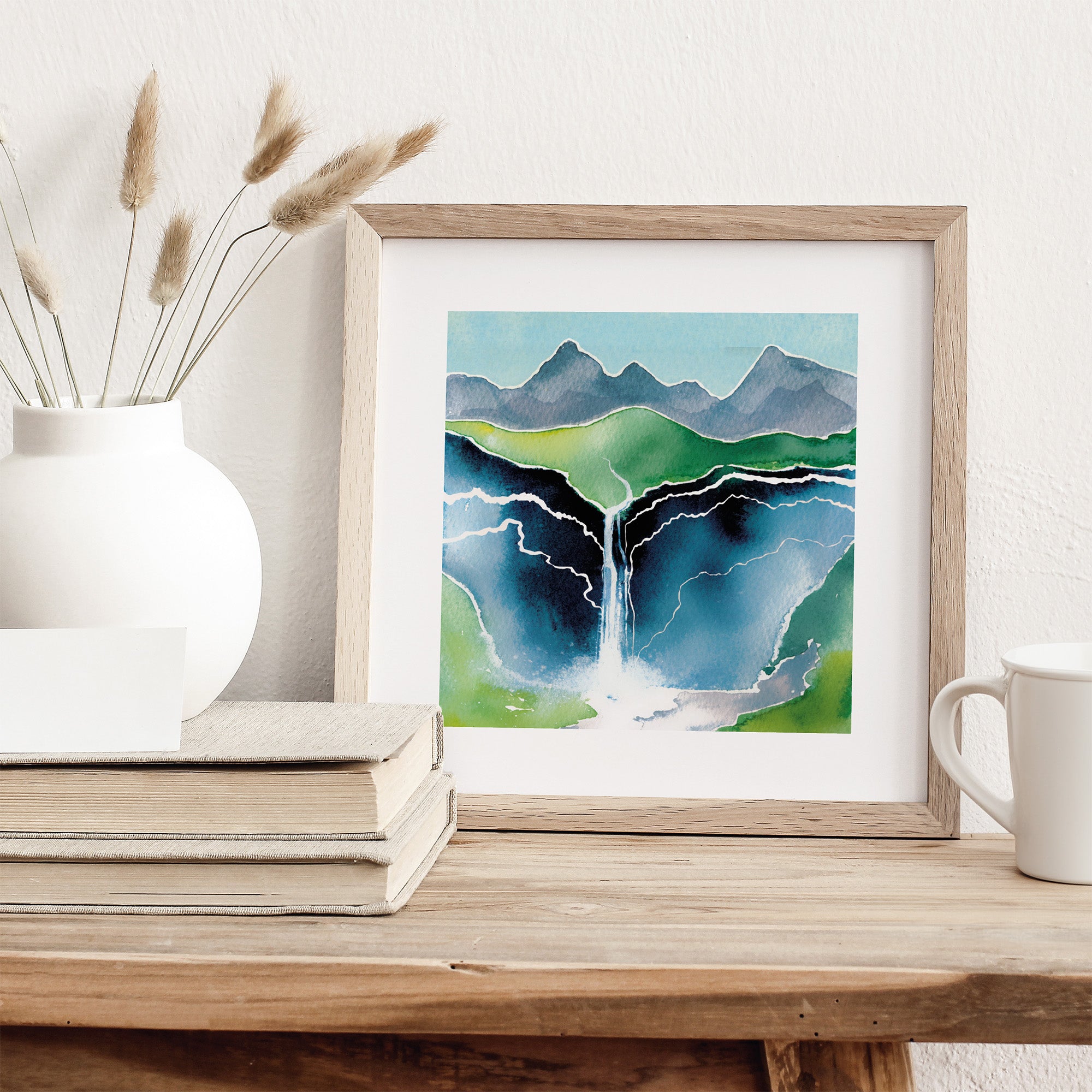 Framed artwork of a mountain landscape on a wooden surface with a vase and mug.
