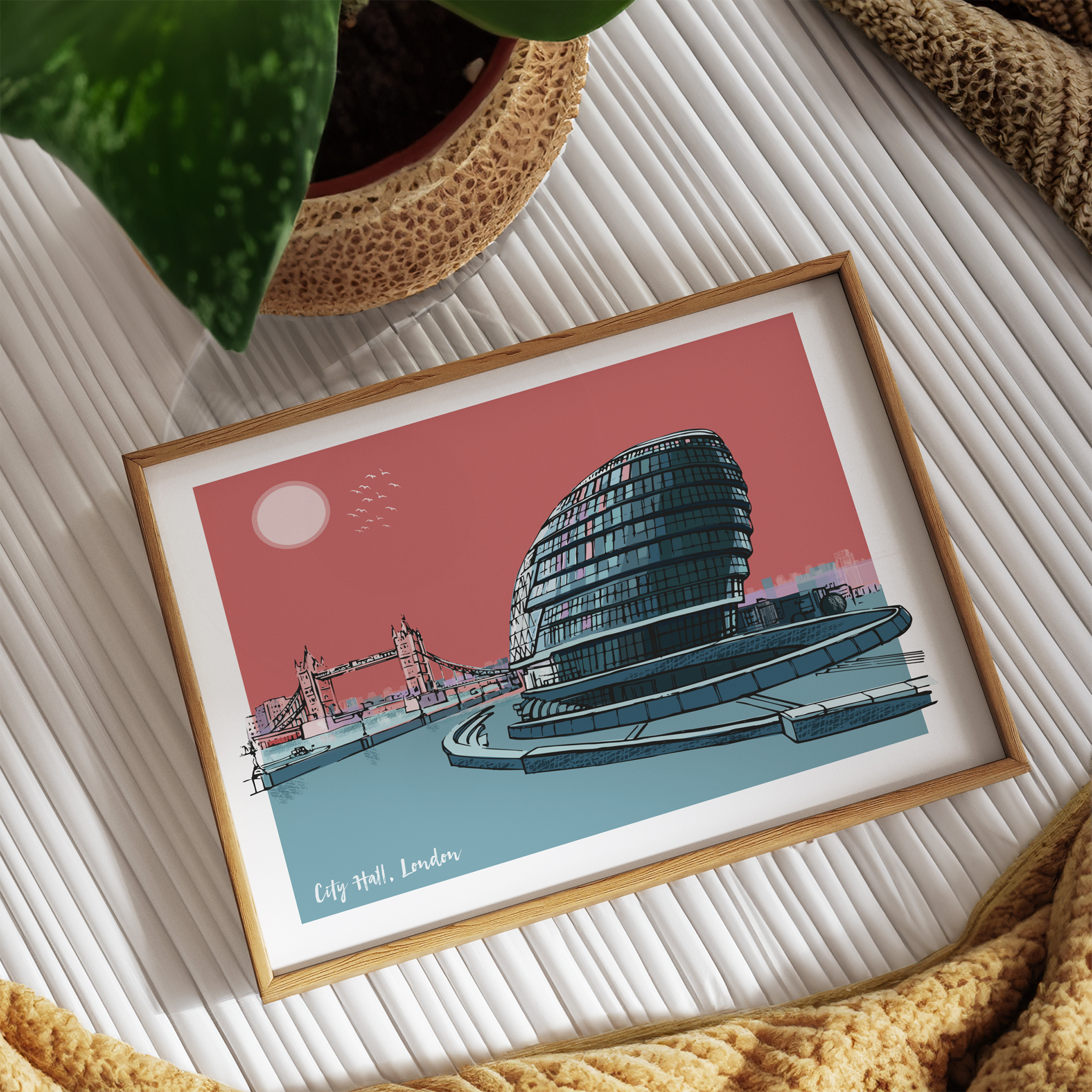 Framed artwork of a building and bridge on a textured surface with a plant and fabric in the background.