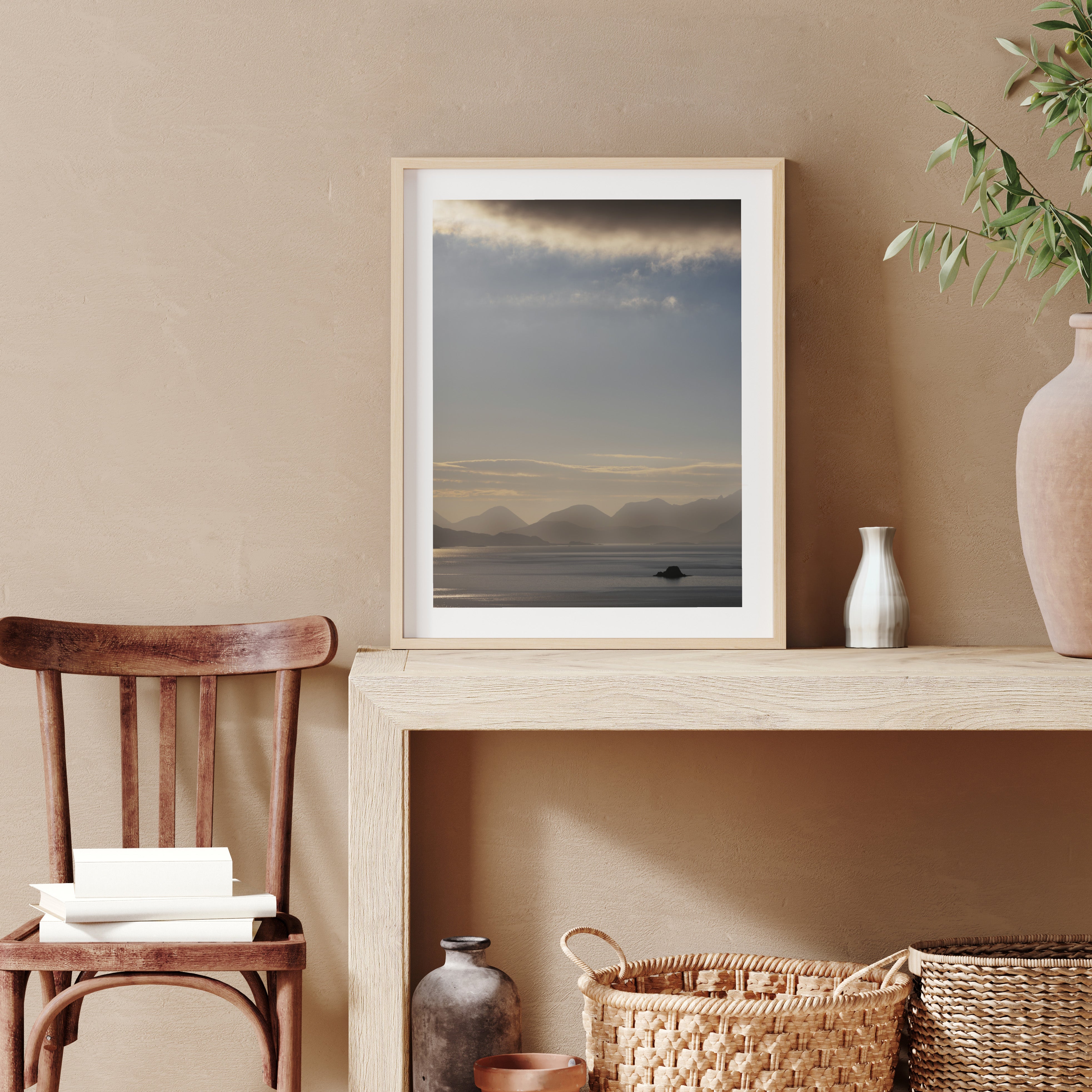 Framed photography art print featuring a sweeping scene of the Isle of Sky, including shimmering water, a warm sunrise and moody cliffs in the distance. Print is leaning on a desk against a wall.