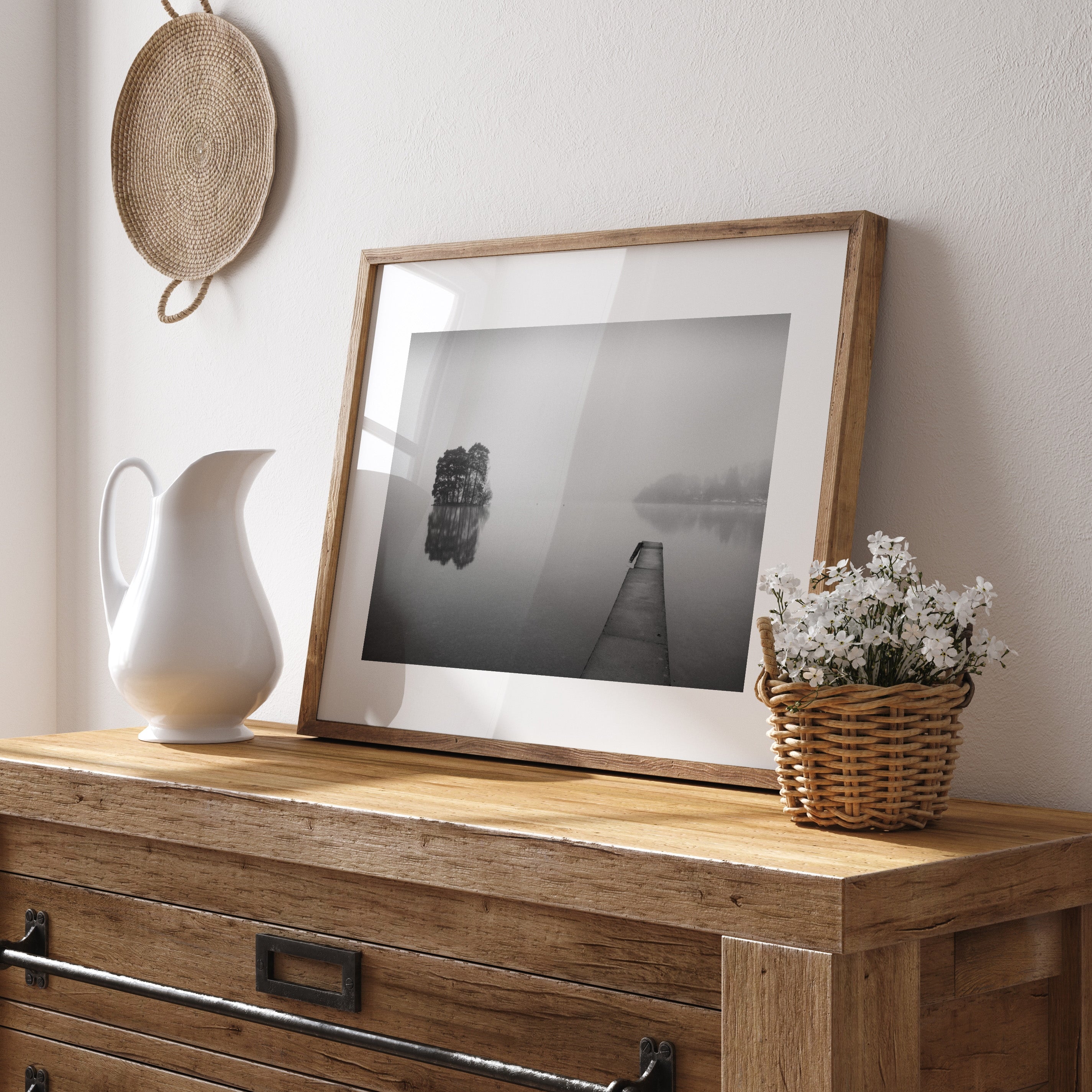 Framed black and white photography art print featuring mist flowing over still waters, perched on a dresser and leaning against a wall.