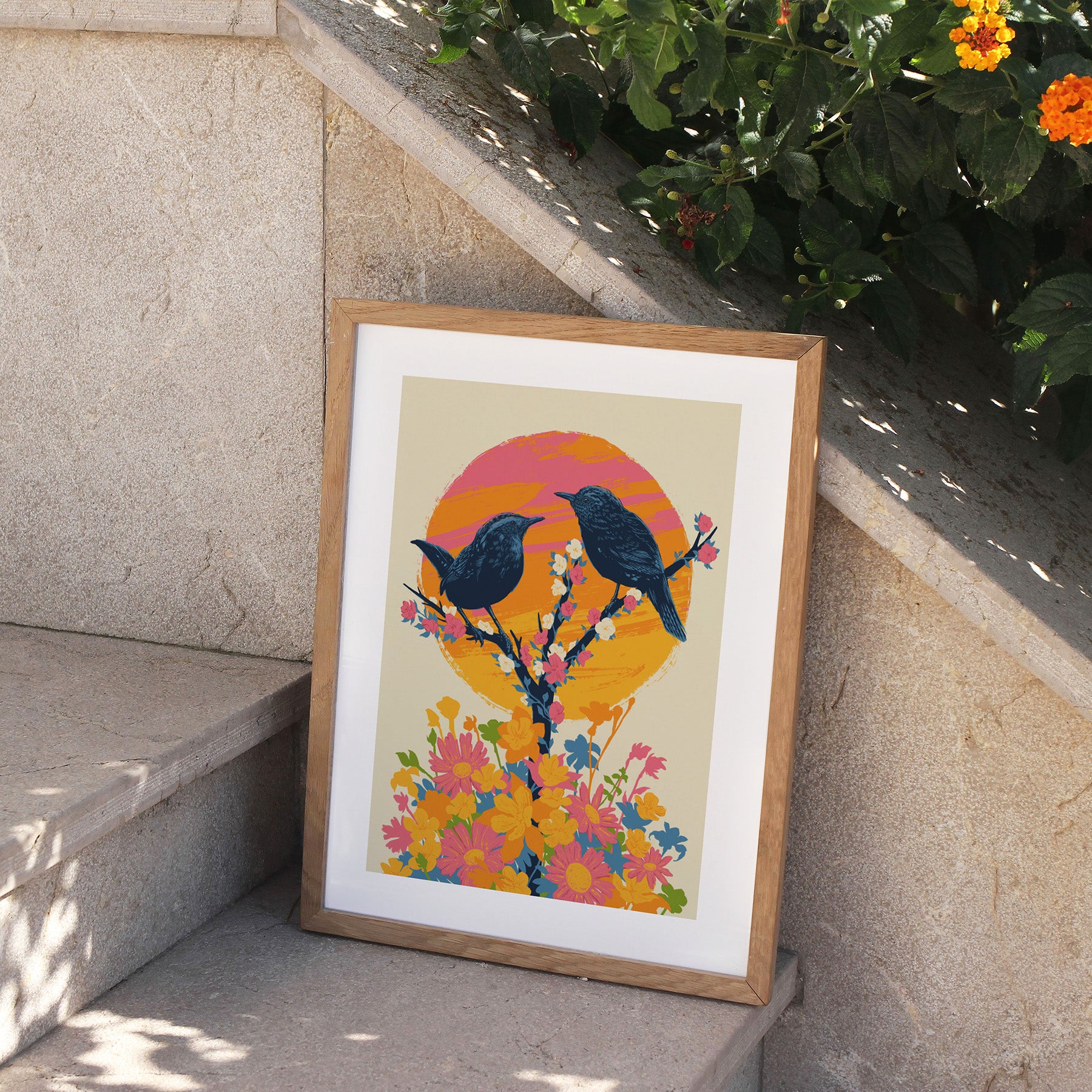 Framed art print featuring a detailed illustration of two birds perching on a blooming tree, in front of a bright sunset. Print is perched on stone steps, outside.