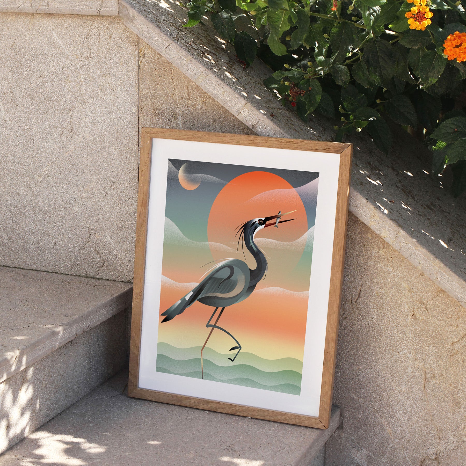 Framed art print featuring a grey heron posing in front of a stunning red sunset by the water. Print is perched on stone steps, outside.