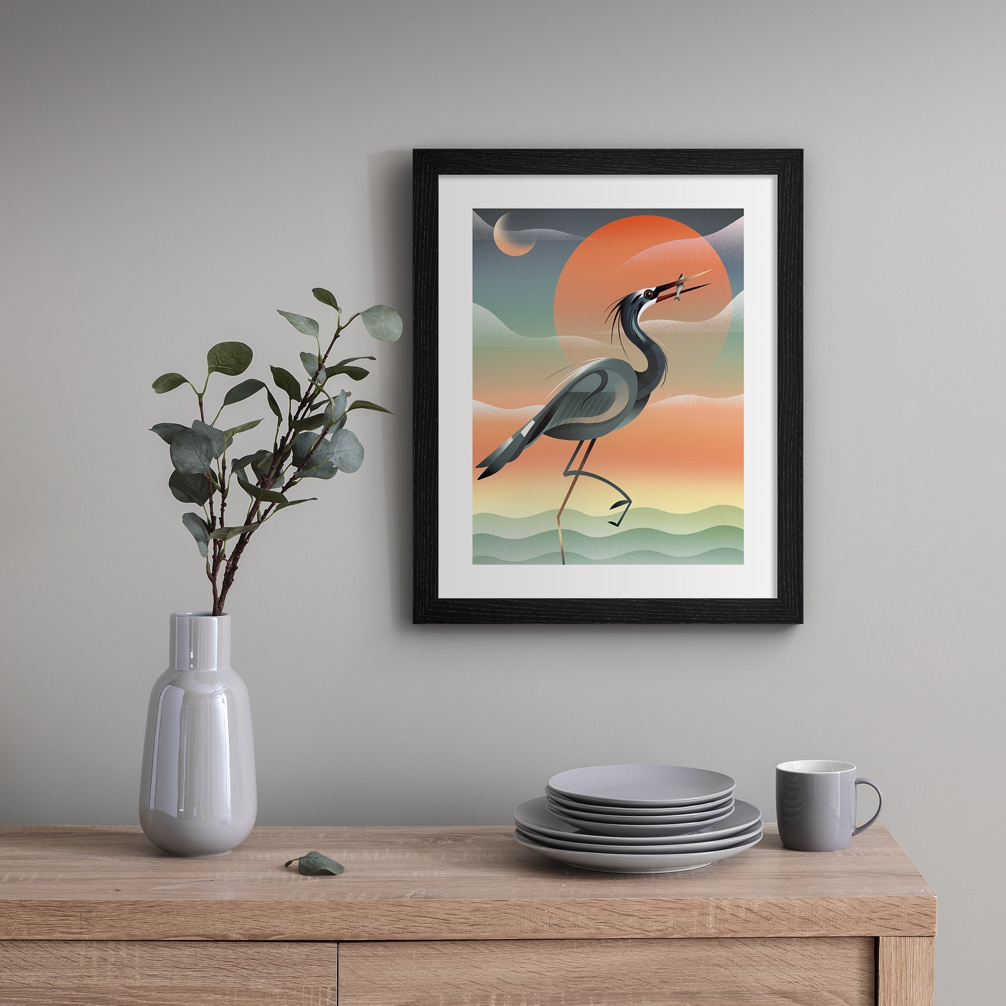 Striking art print featuring a grey heron posing in front of a stunning red sunset by the water. Art print is hung up on a grey wall.