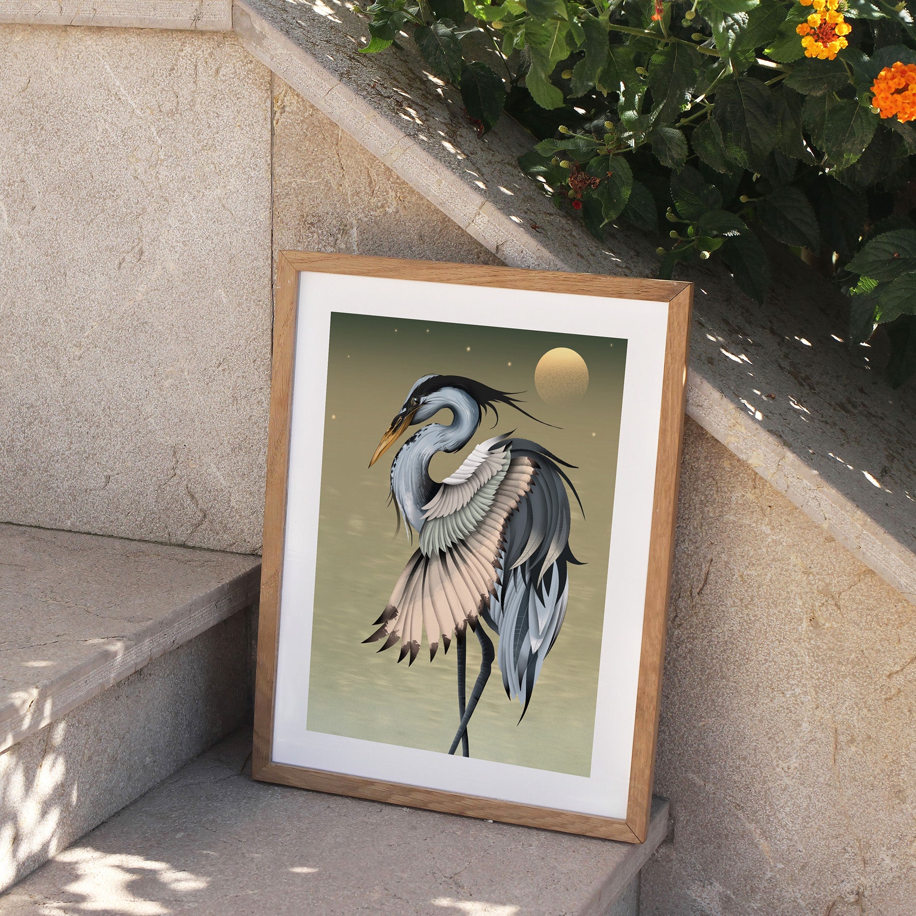 Framed art print featuring a heron basking in front of a golden sun. Print is perched on stone steps, outside.