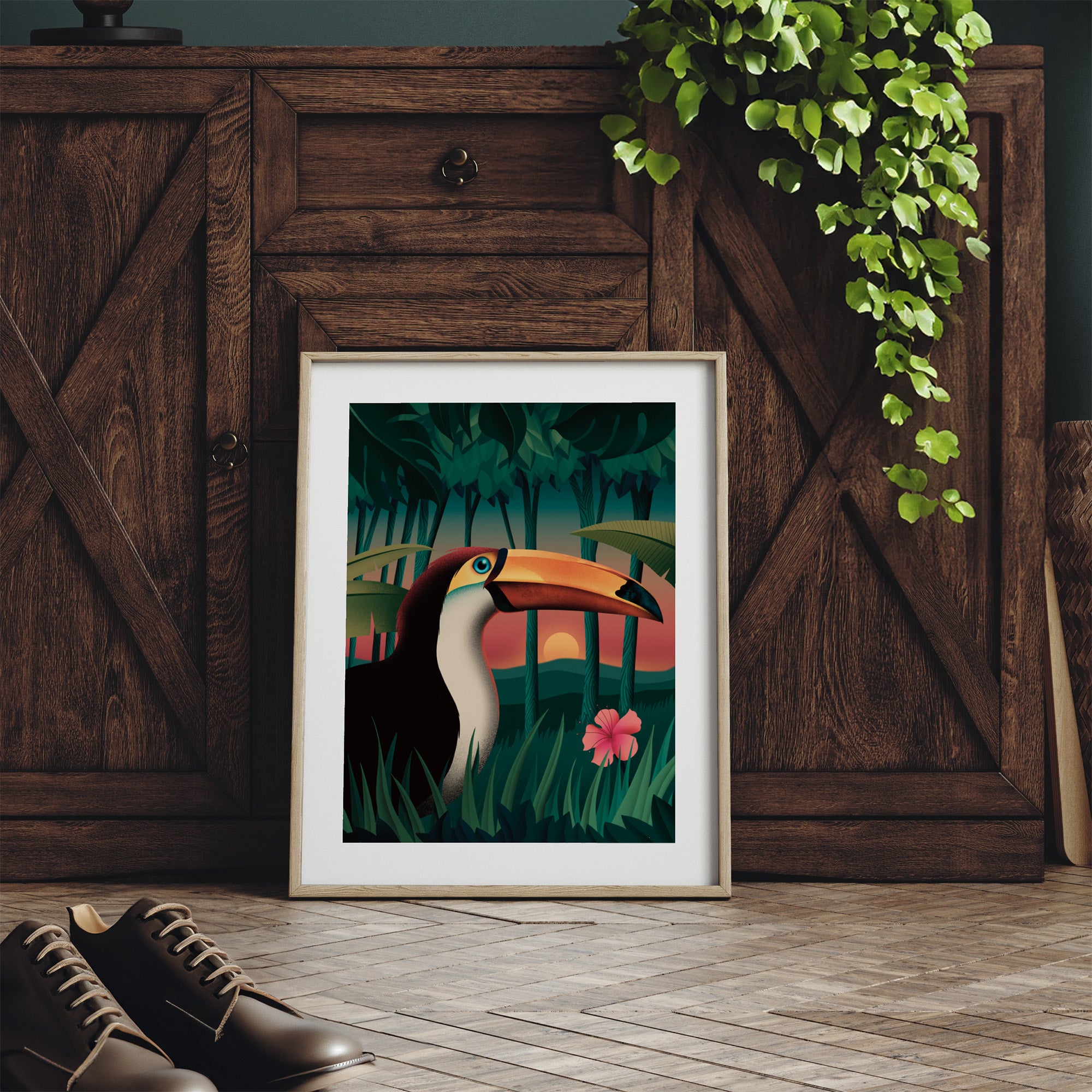 Framed art print featuring a sunset forest scene of a toucan standing amidst the trees. Print is leaning against a sideboard table.