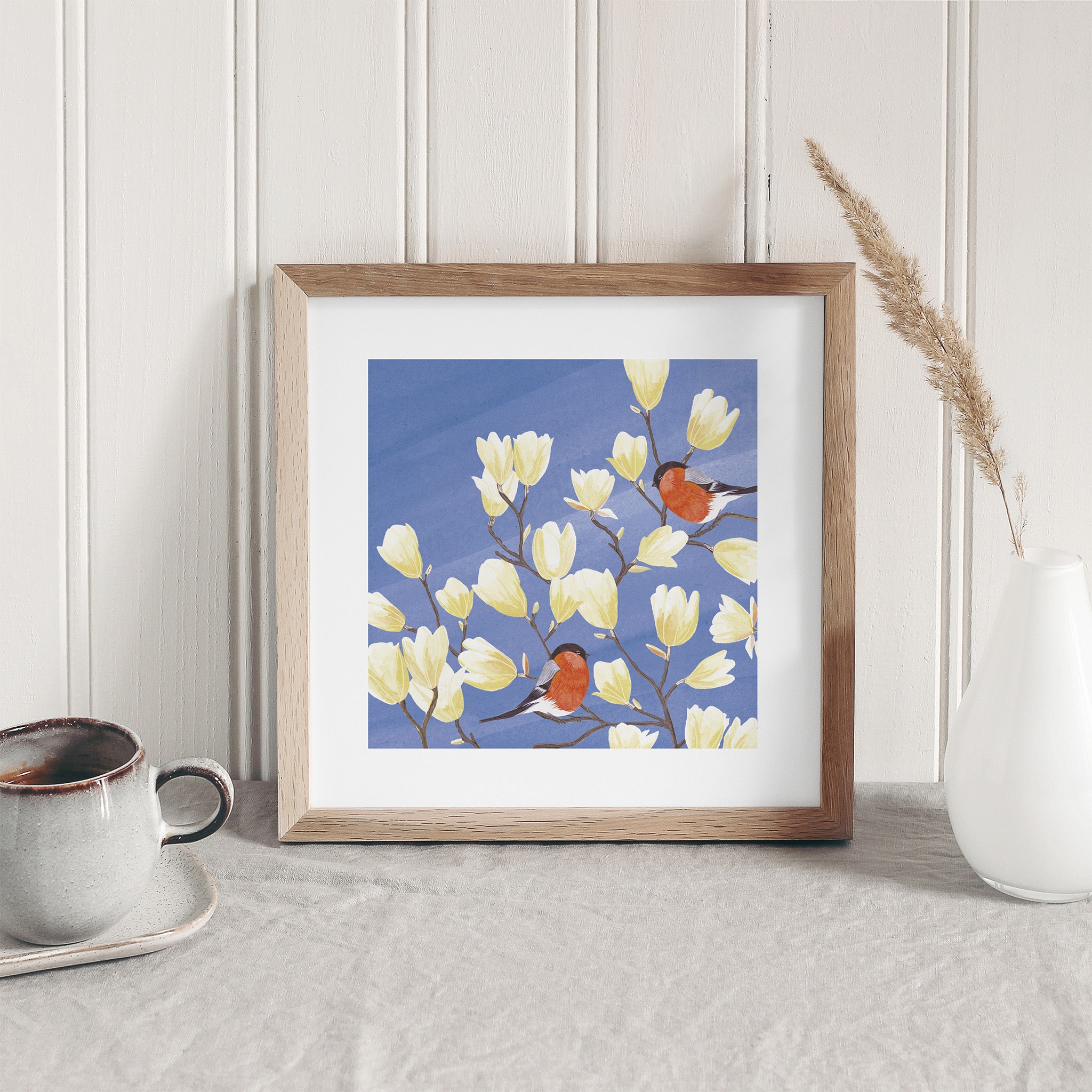 Framed artwork of birds and flowers on a blue background, placed on a surface with a cup and vase.
