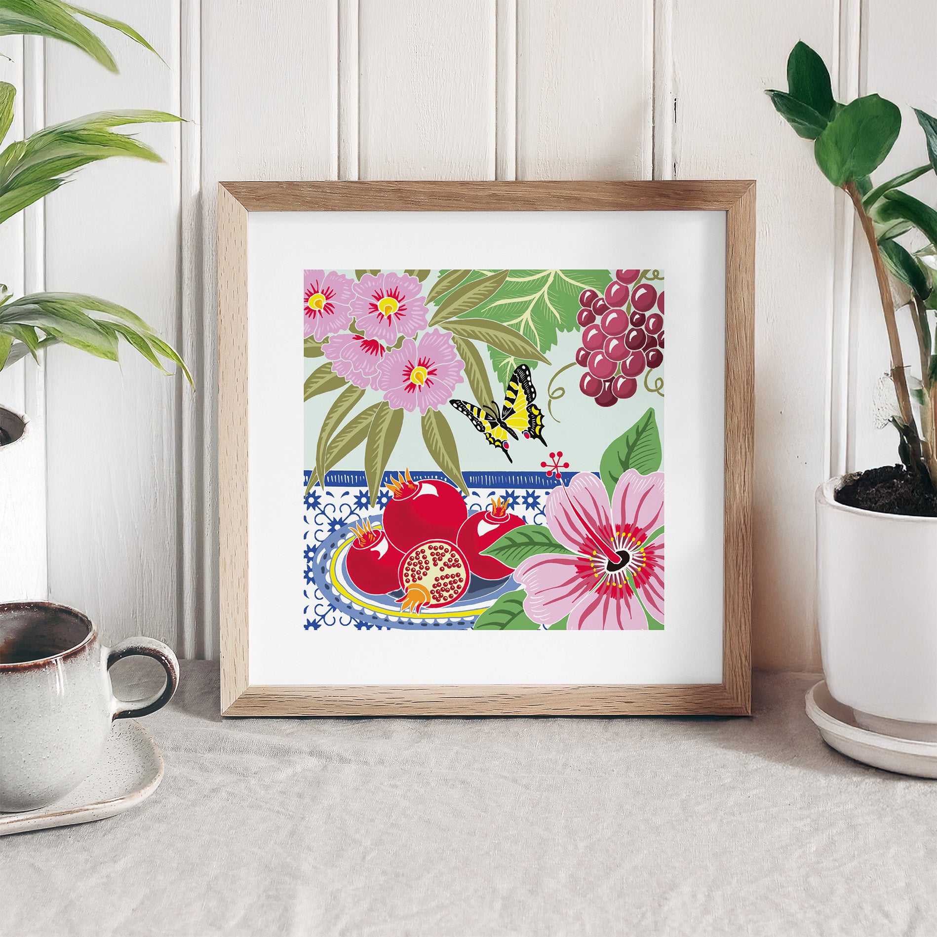 Framed artwork with floral and butterfly design on a white surface with plants and a cup.