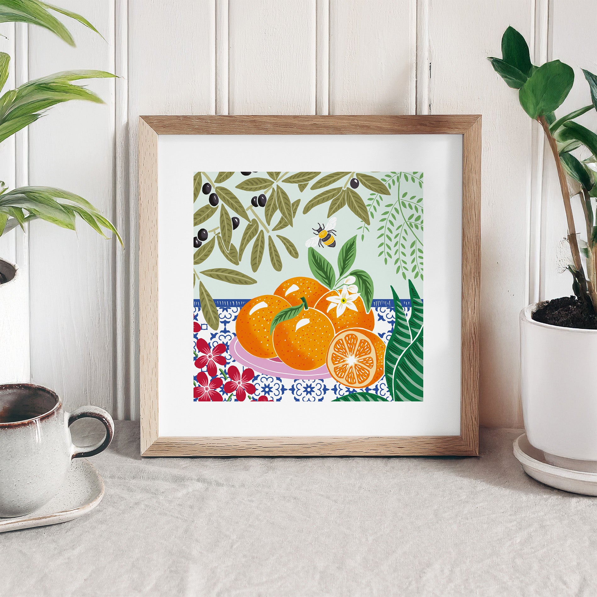 Framed artwork with oranges and floral patterns on a white surface with plants and a cup.