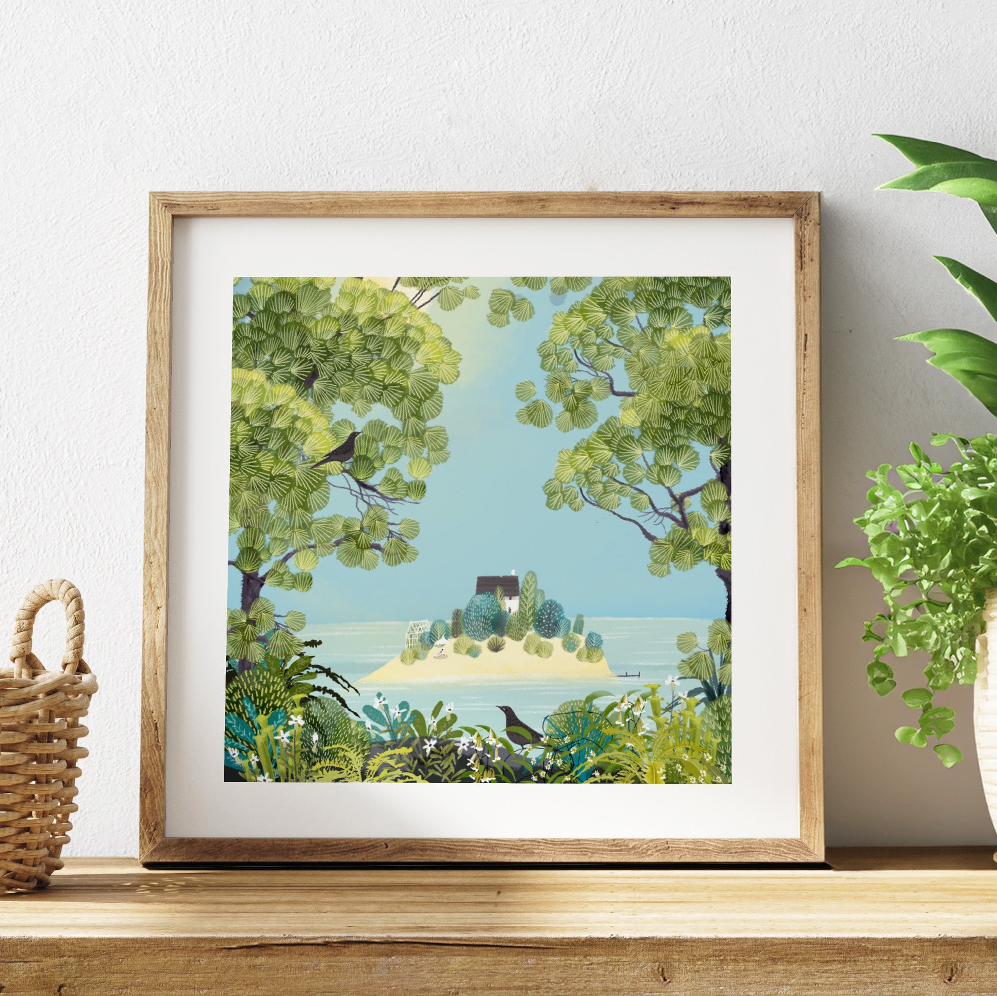 Framed artwork of a scenic landscape with trees and a house on an island, placed on a wooden surface.