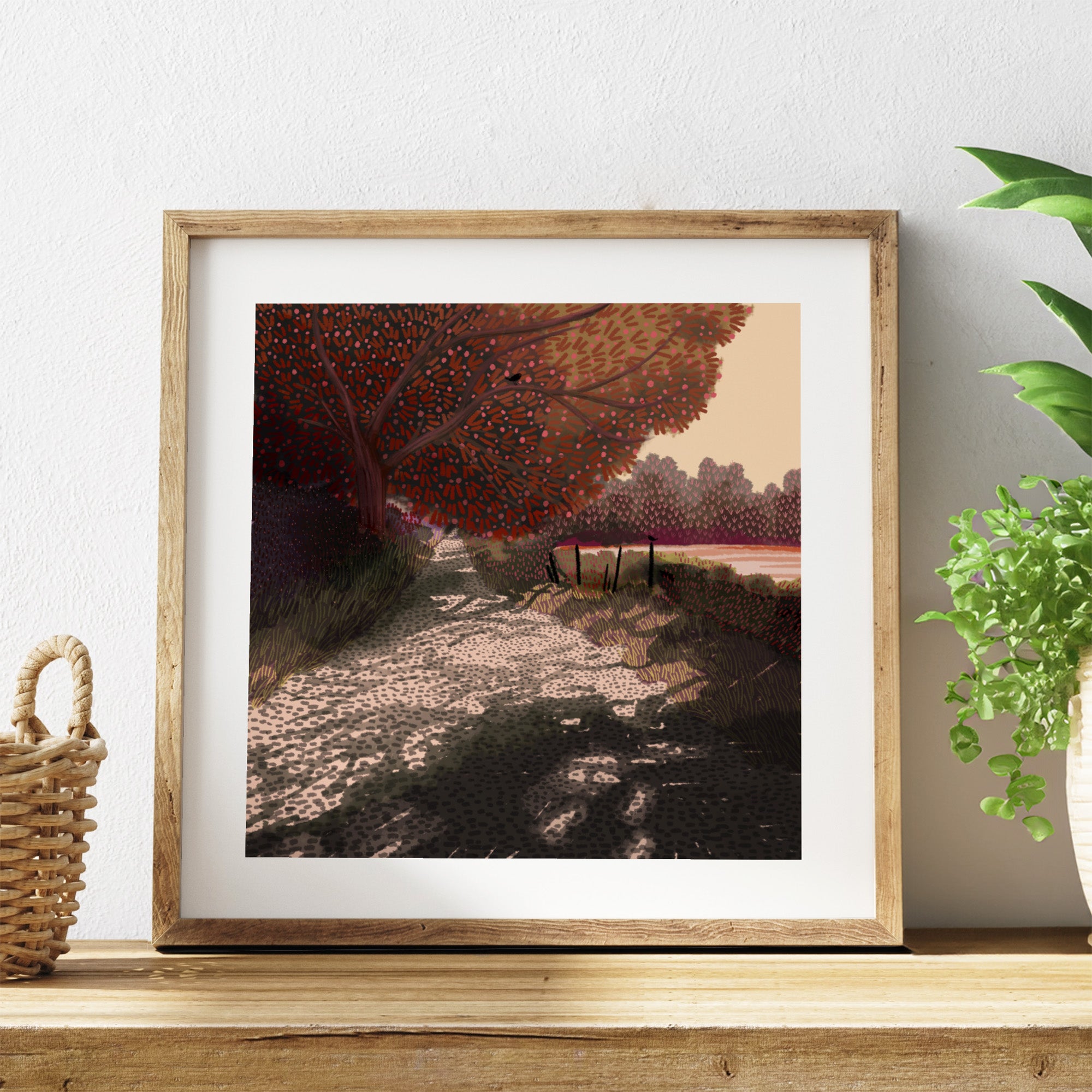 Framed artwork of an autumn landscape with a path and trees on a white wall.